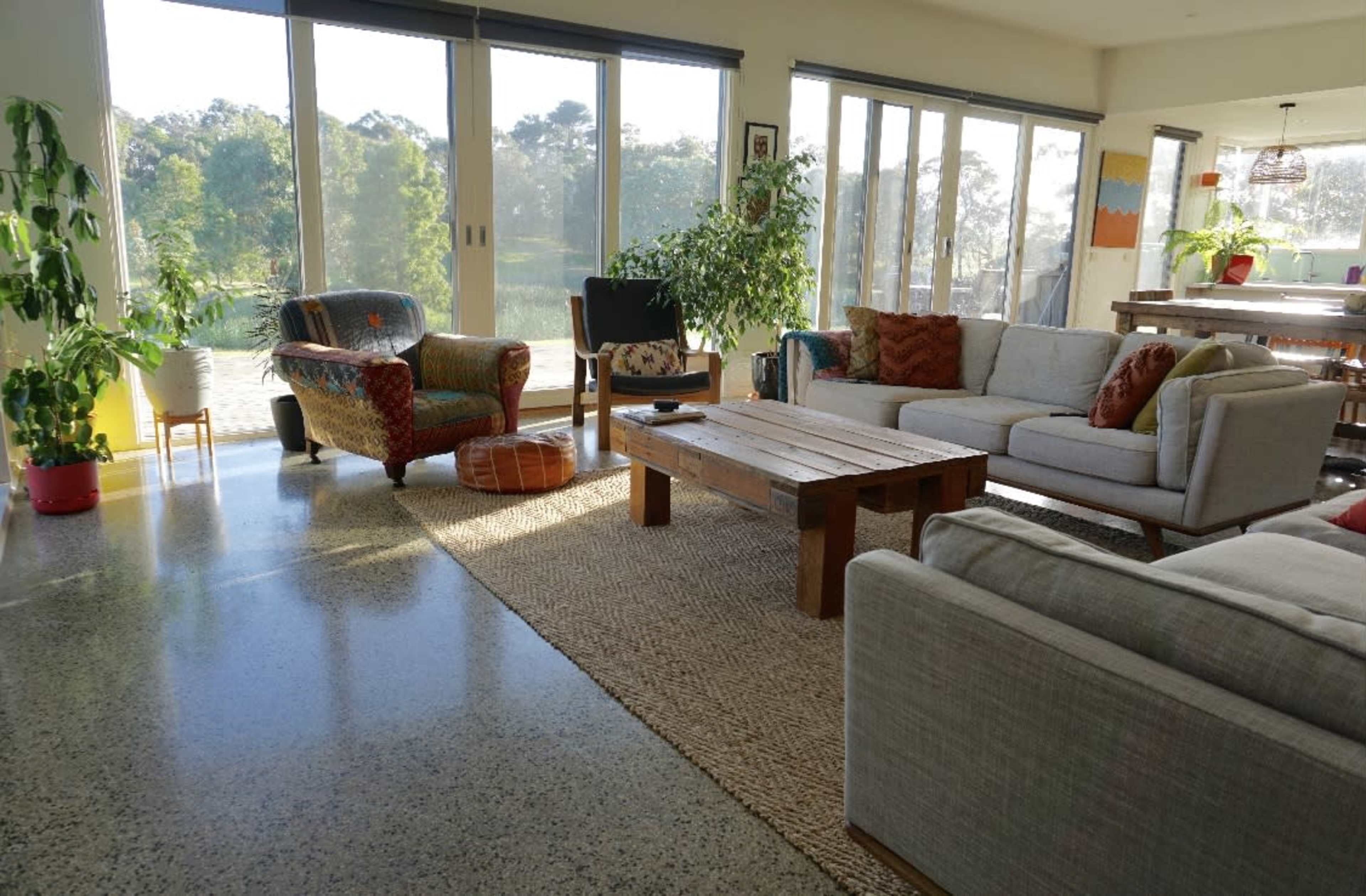 A spacious living room features large windows with views of greenery, comfortable seating, and a wooden coffee table on a textured rug.