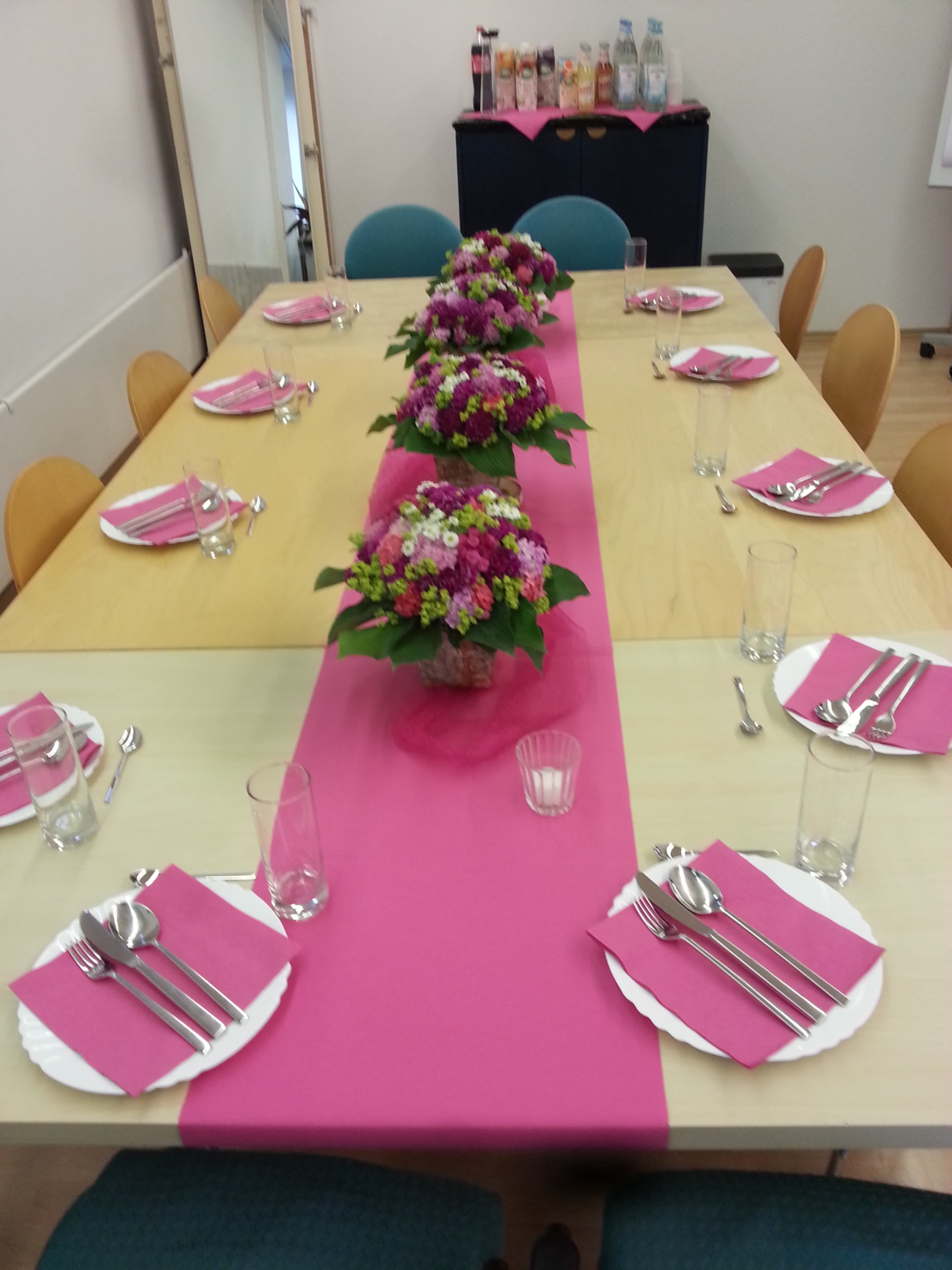 A long dining table is set with plates, utensils, and glasses, adorned with vibrant floral centerpieces and a pink table runner.