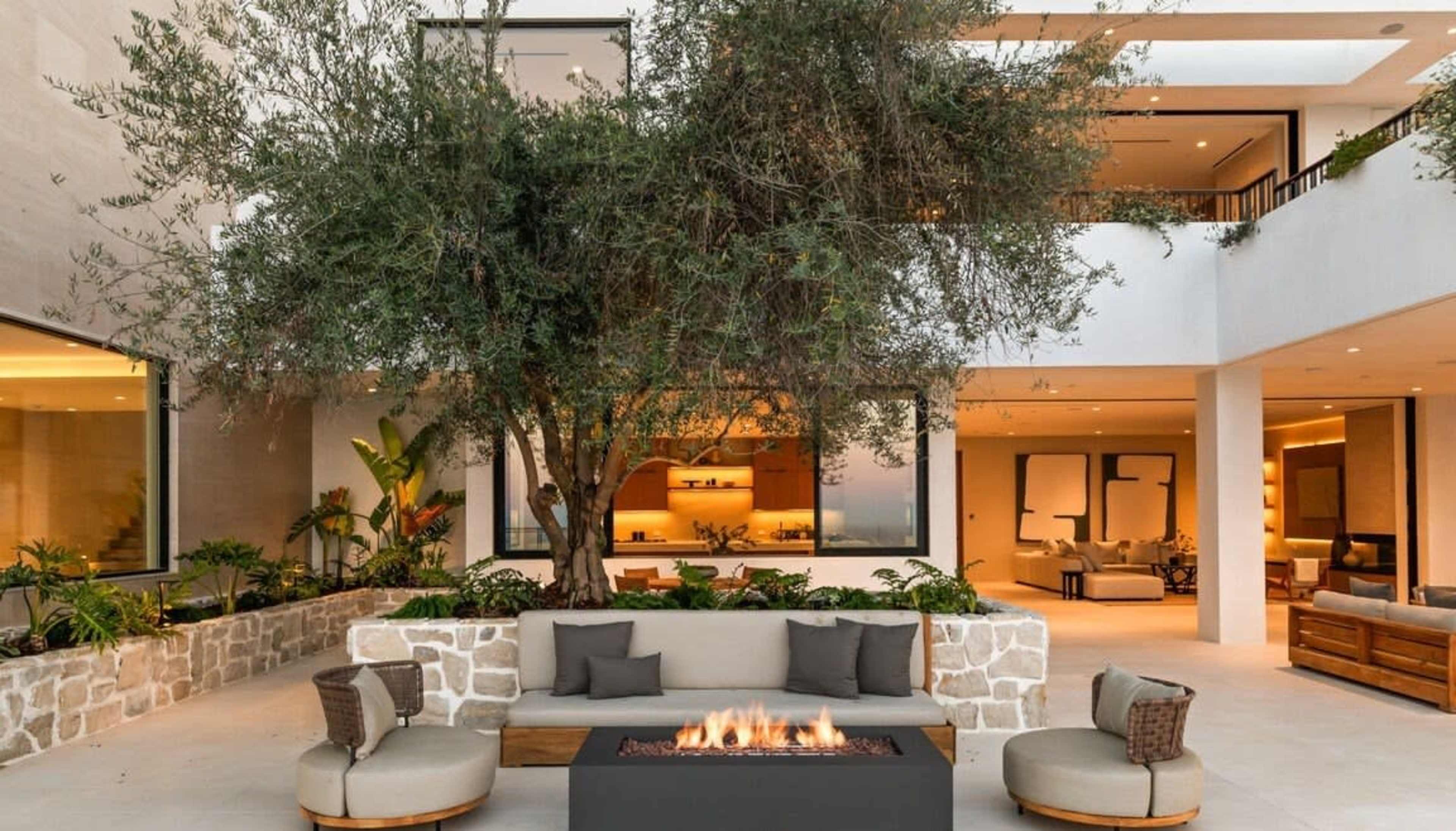 The image shows a modern outdoor seating area with a fire pit surrounded by a large olive tree and various plants.