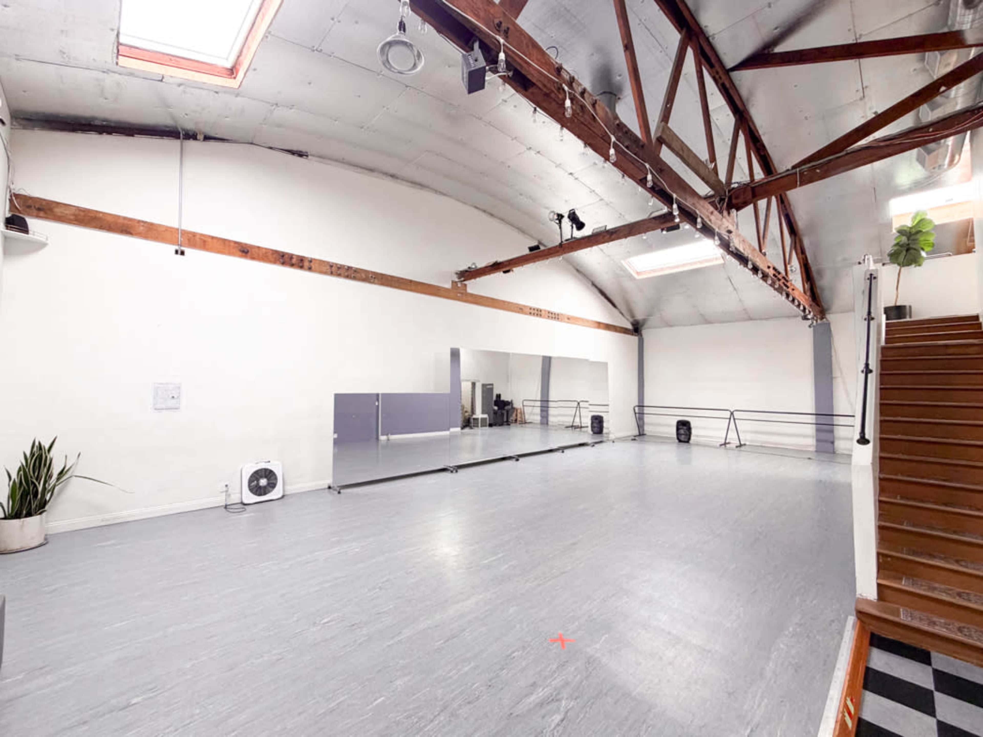 The image shows a spacious studio with a wooden beam ceiling, large mirrors, and wooden stairs leading up to the entrance.