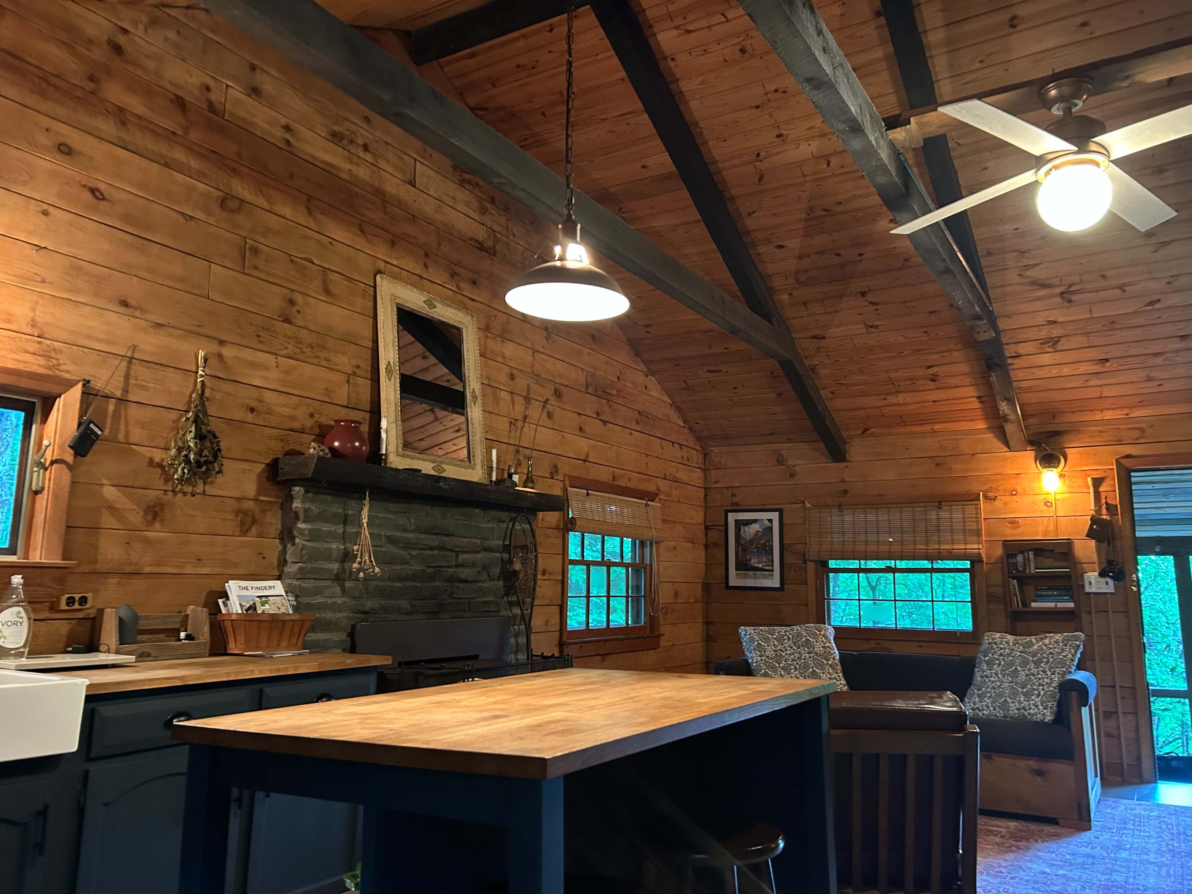 The image shows a wooden cabin interior featuring a kitchen island, a stone fireplace, and a cozy seating area with large windows.