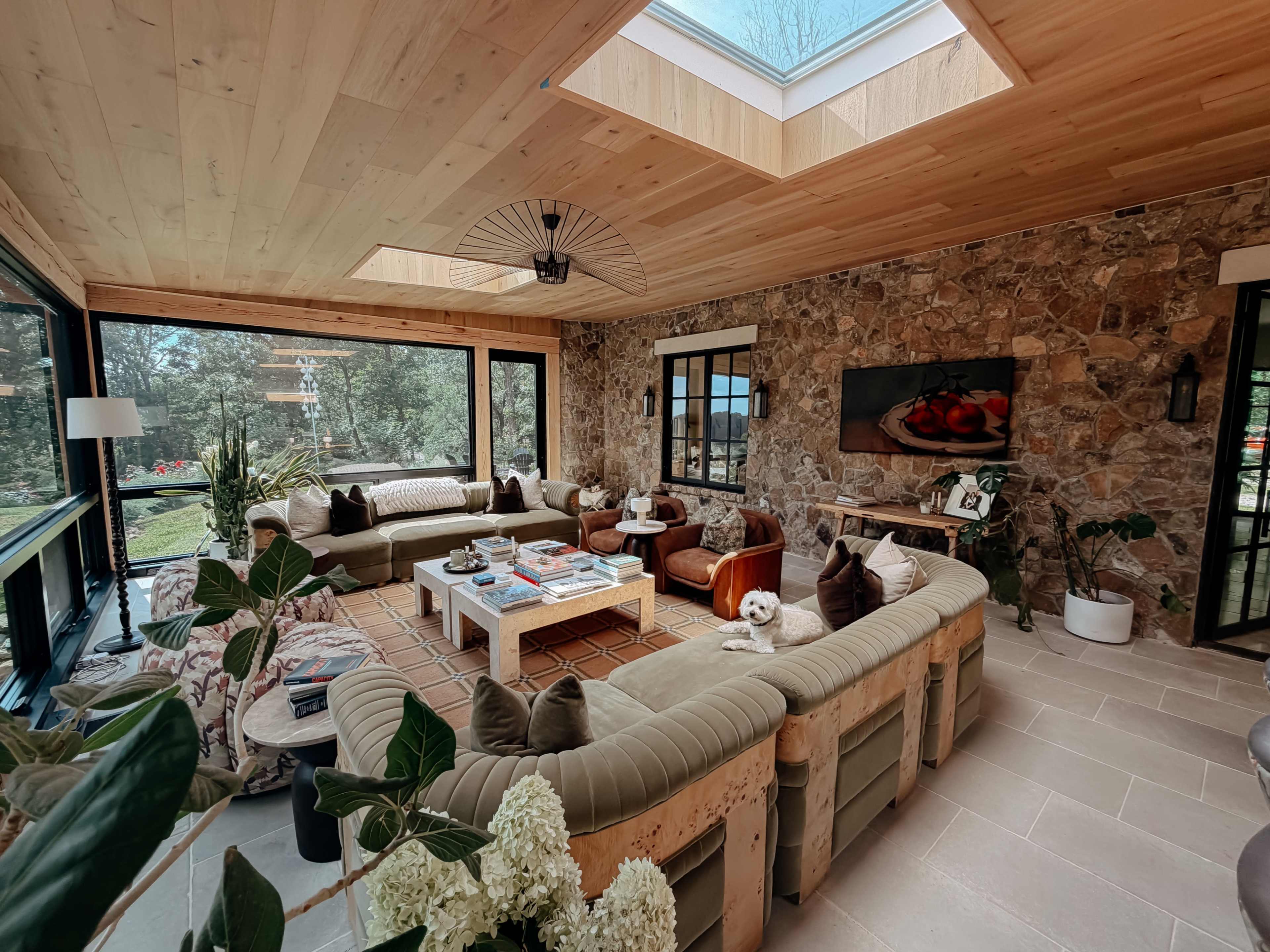 The image shows a cozy sunroom featuring a circular sectional sofa, large windows with views of trees, a skylight, and a stone wall accent.