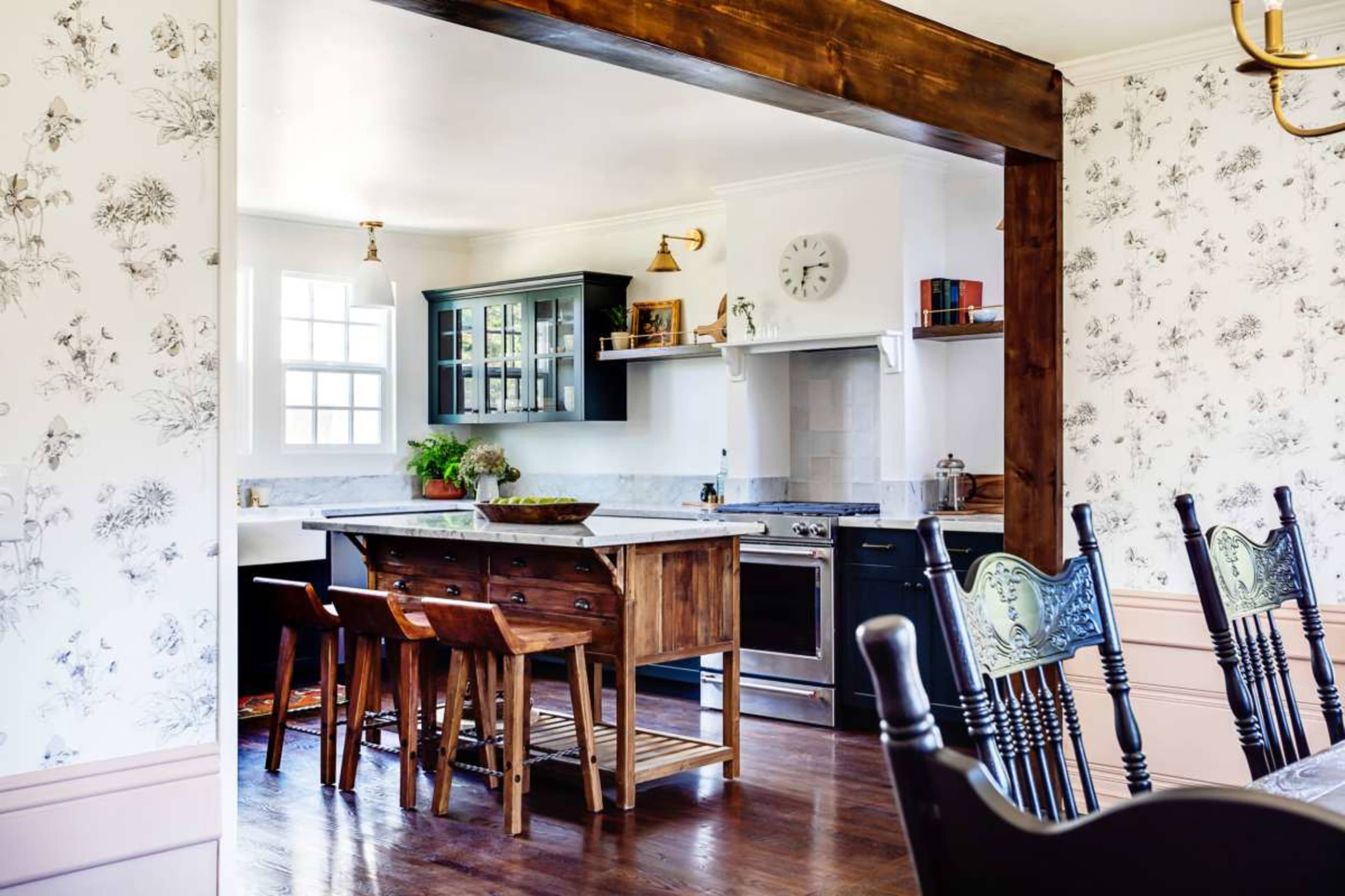 A modern kitchen features a central island with wooden stools, dark cabinetry, and floral wallpaper.