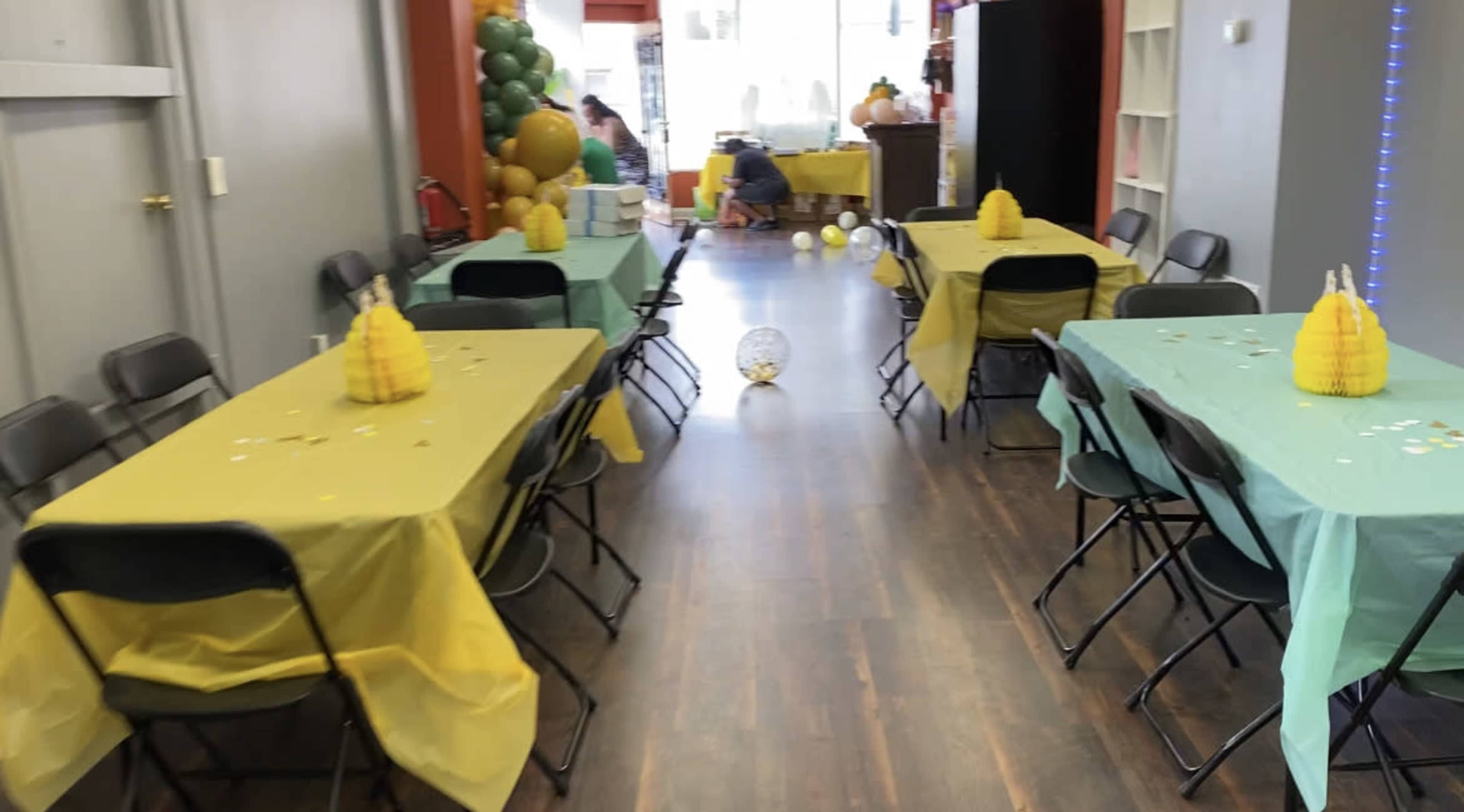 A party venue with tables covered in yellow and green tablecloths, decorated with pineapple centerpieces, and balloons arranged along the walls.