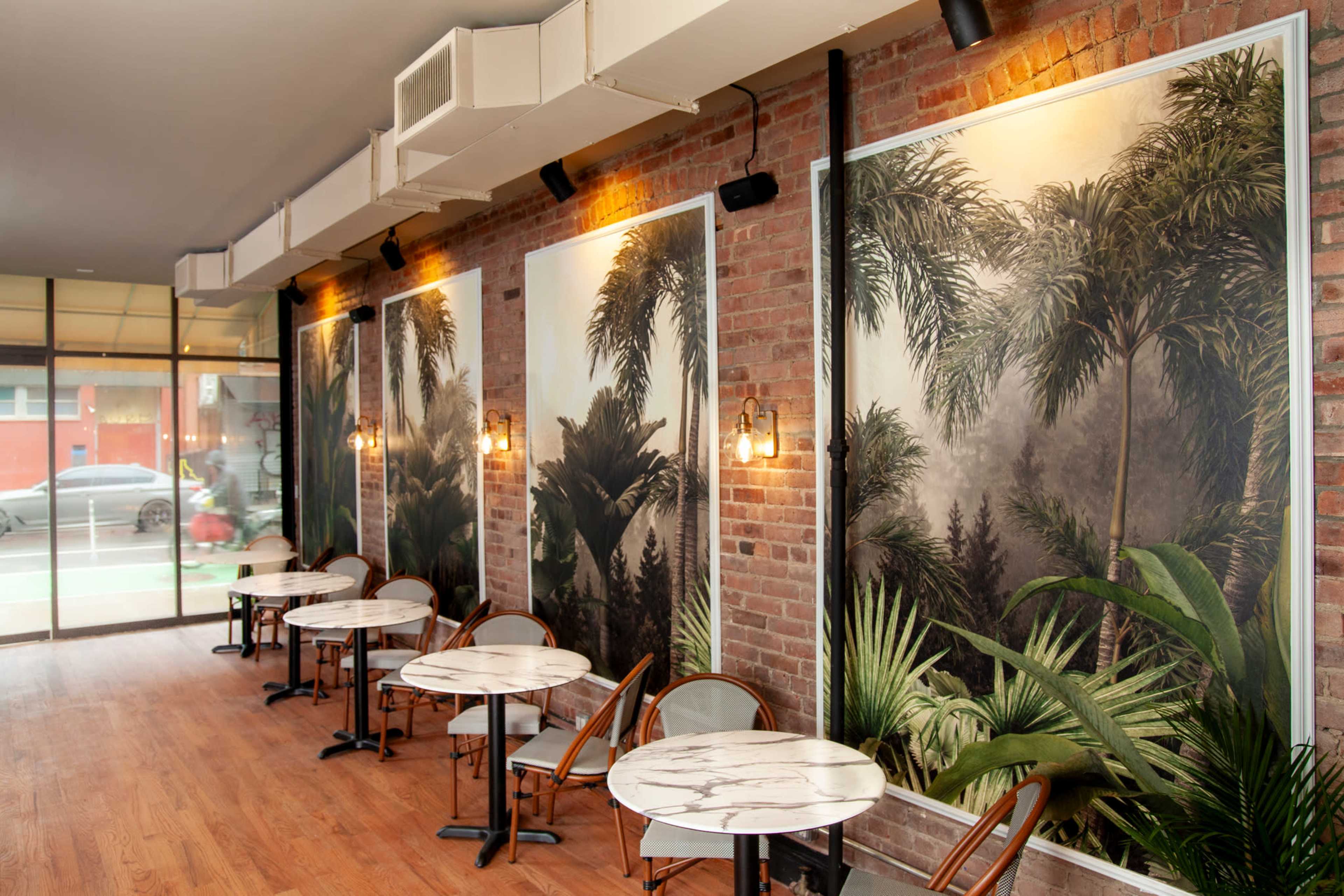 A cozy café with brick walls adorned by large tropical-themed murals, featuring round marble tables and chairs arranged neatly.