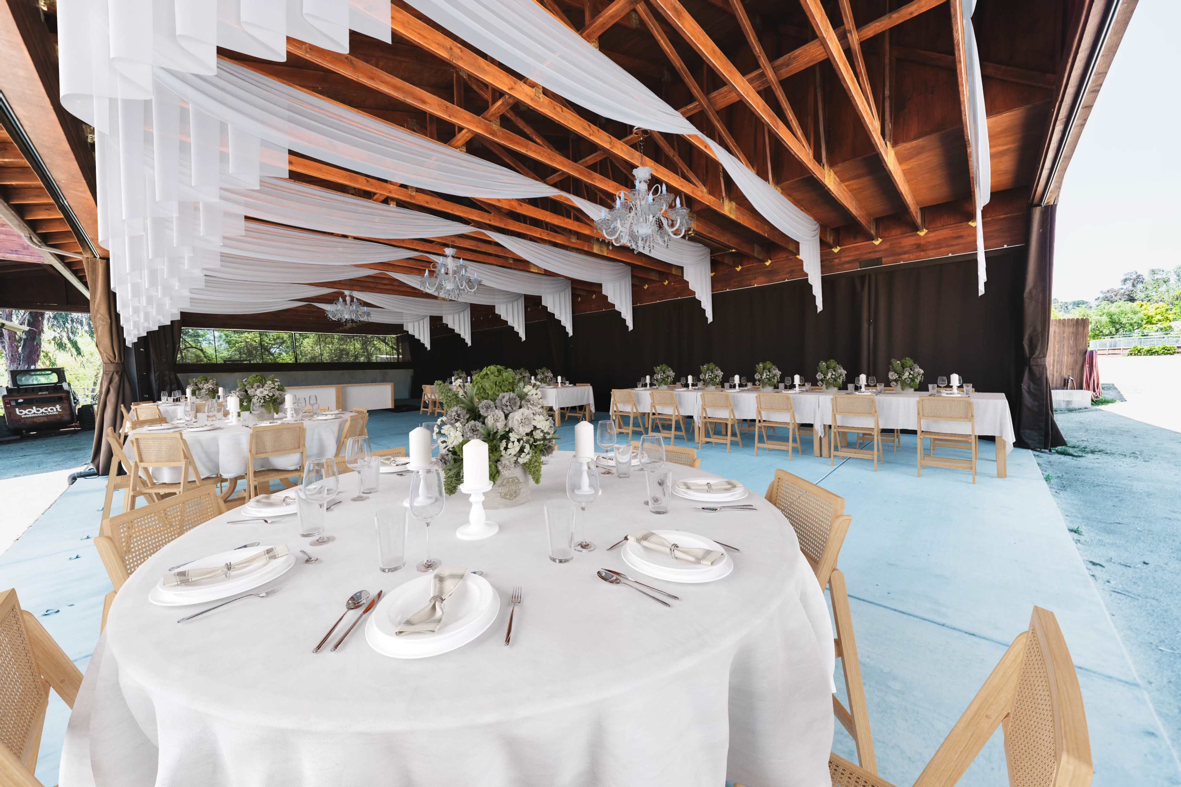A spacious outdoor dining area features multiple tables dressed with white linens under decorative drapery and chandeliers.