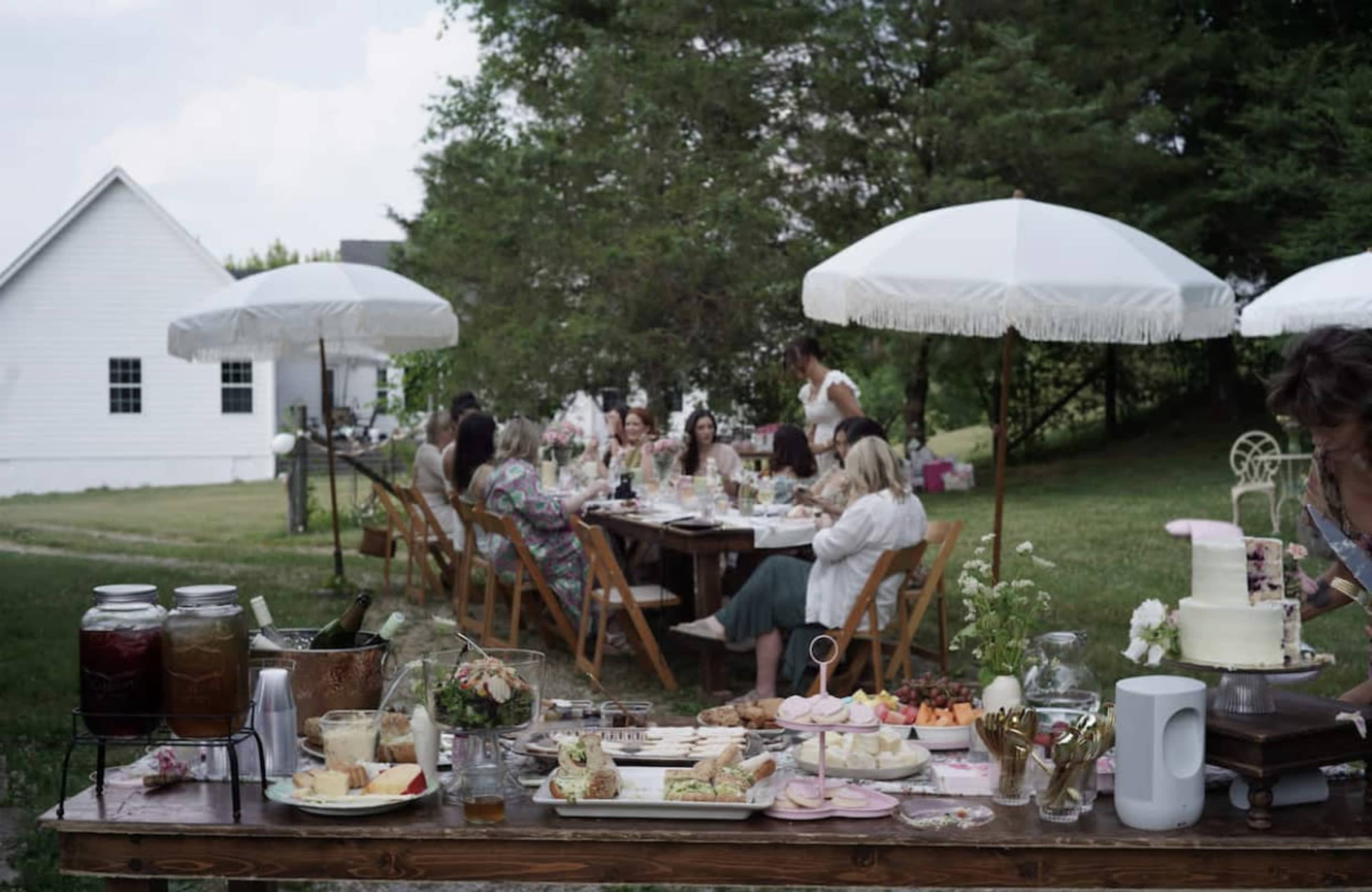 A group of people gathered at a long table outdoors, enjoying a meal under white umbrellas in a garden setting with a house in the background.