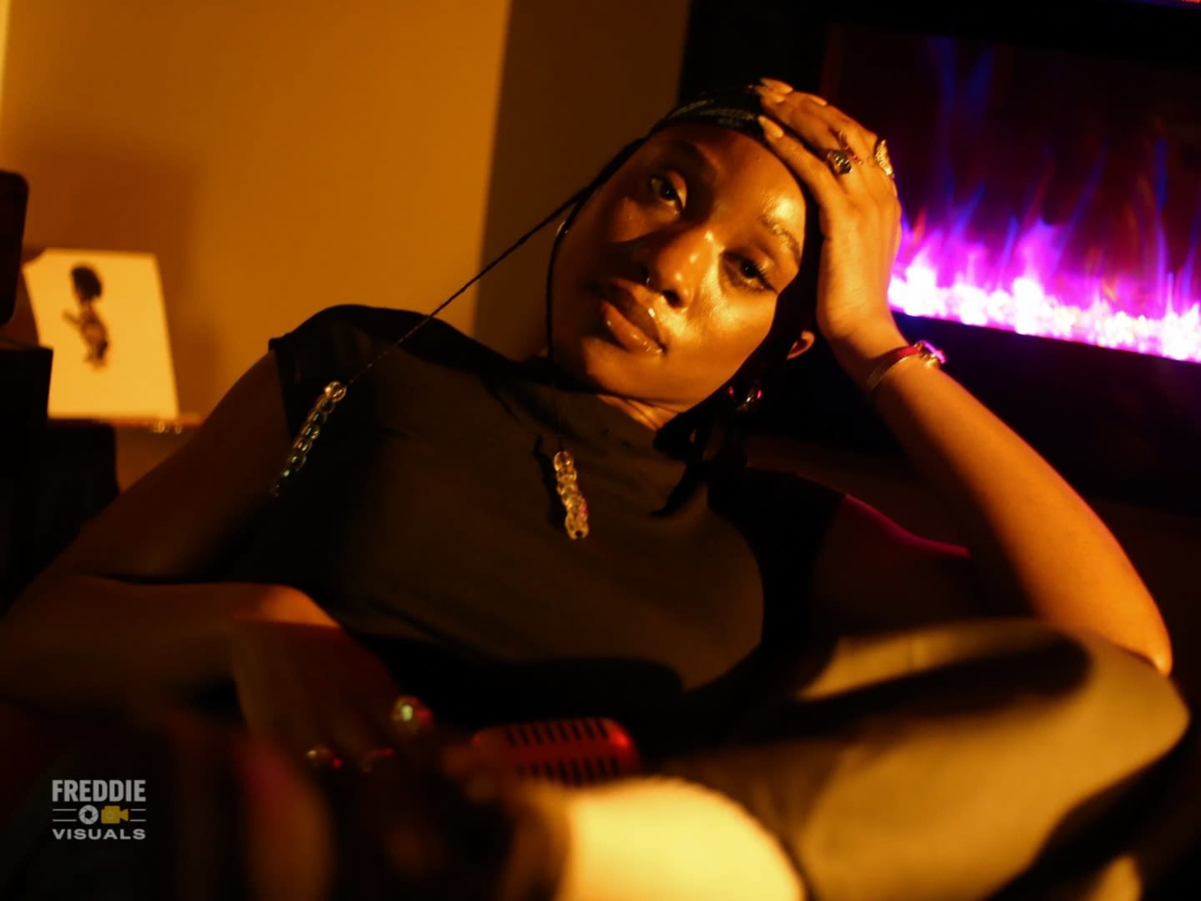 A person is sitting on a couch with their hand on their head, while a glowing fireplace is visible in the background.