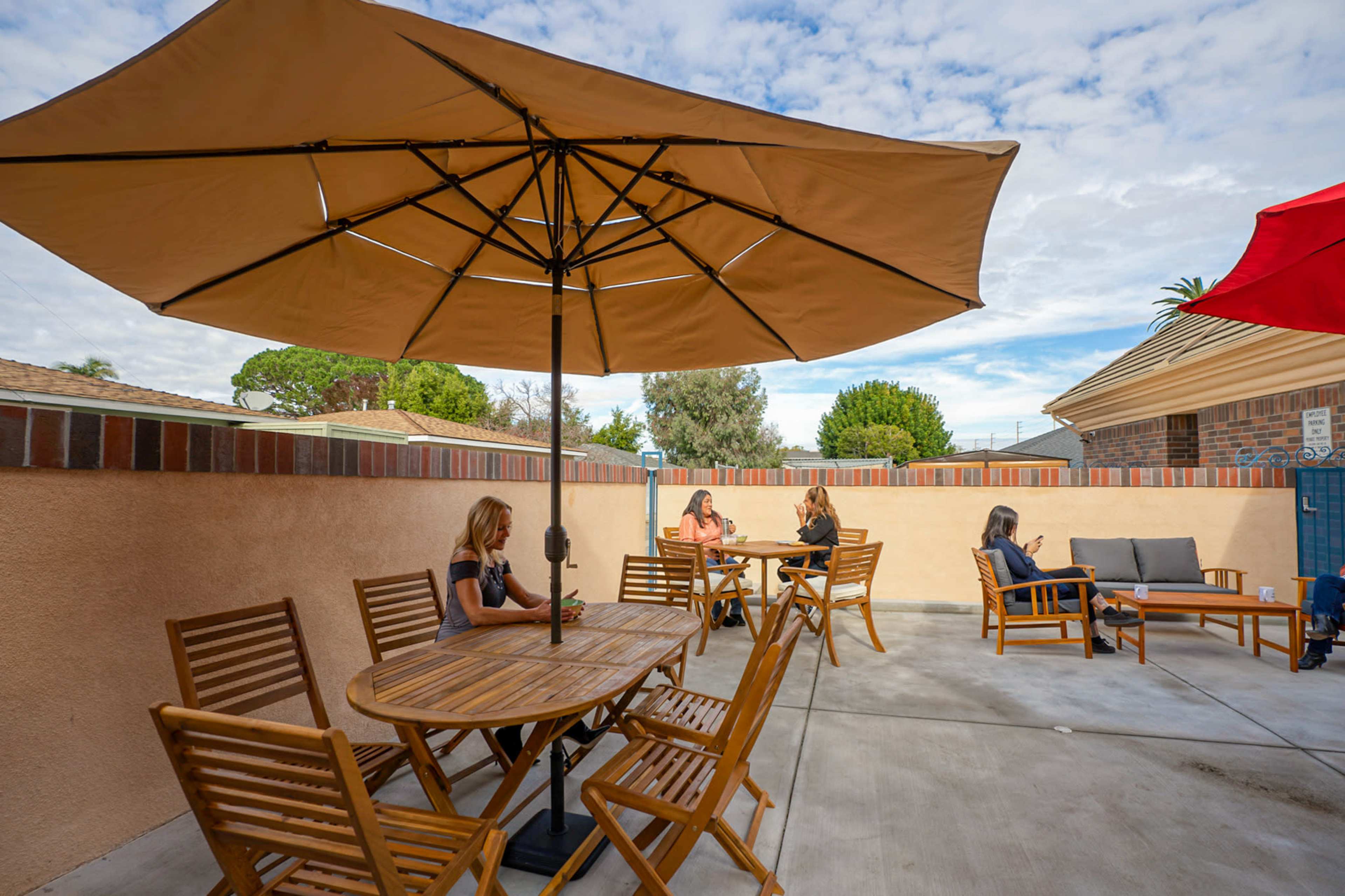 A group of four people sit at wooden tables under umbrellas in an outdoor seating area surrounded by a low wall.