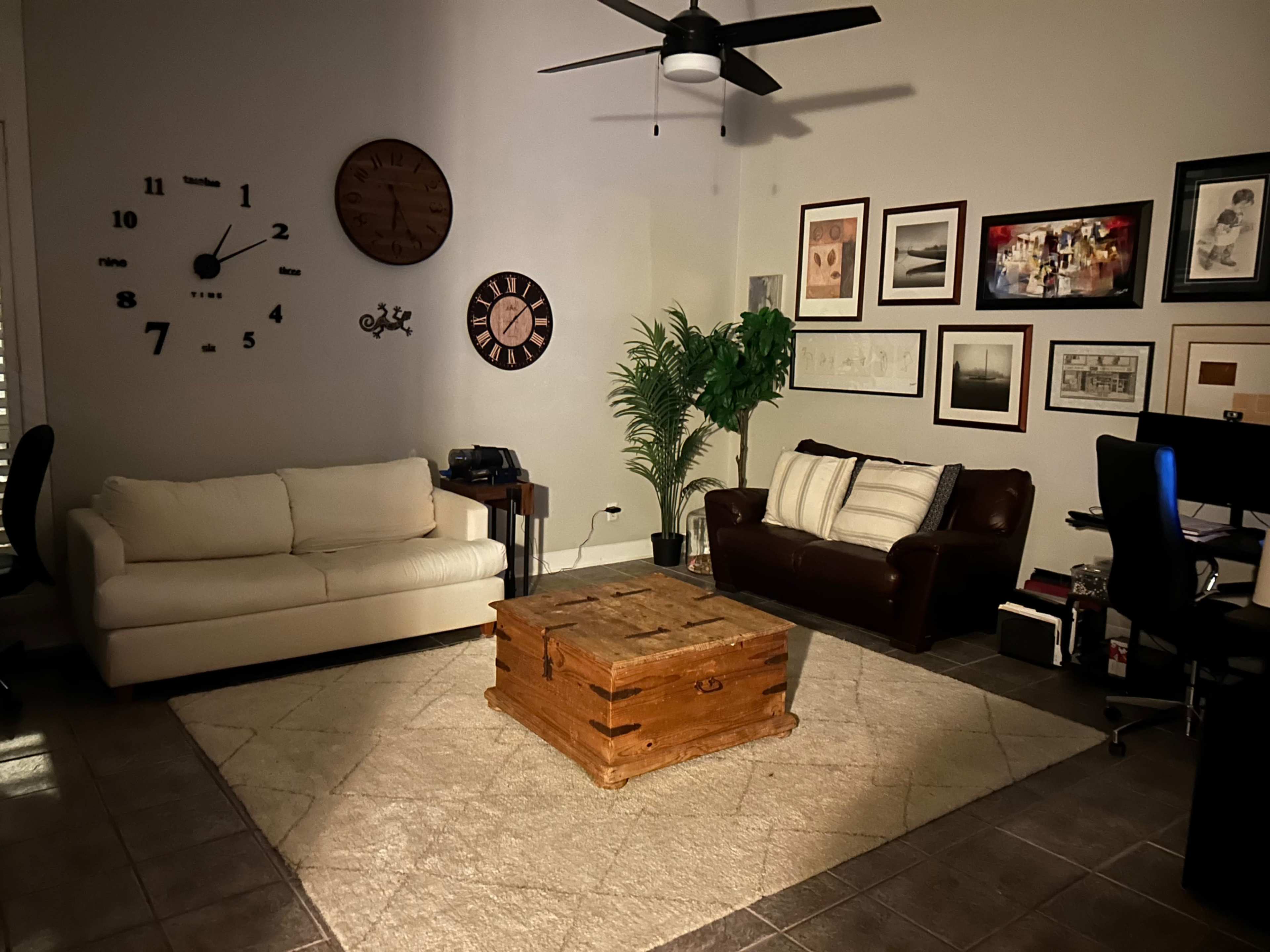 The image shows a living room featuring a white sofa, a brown leather couch, a large wooden coffee table, and various framed pictures on the walls, with a ceiling fan overhead.
