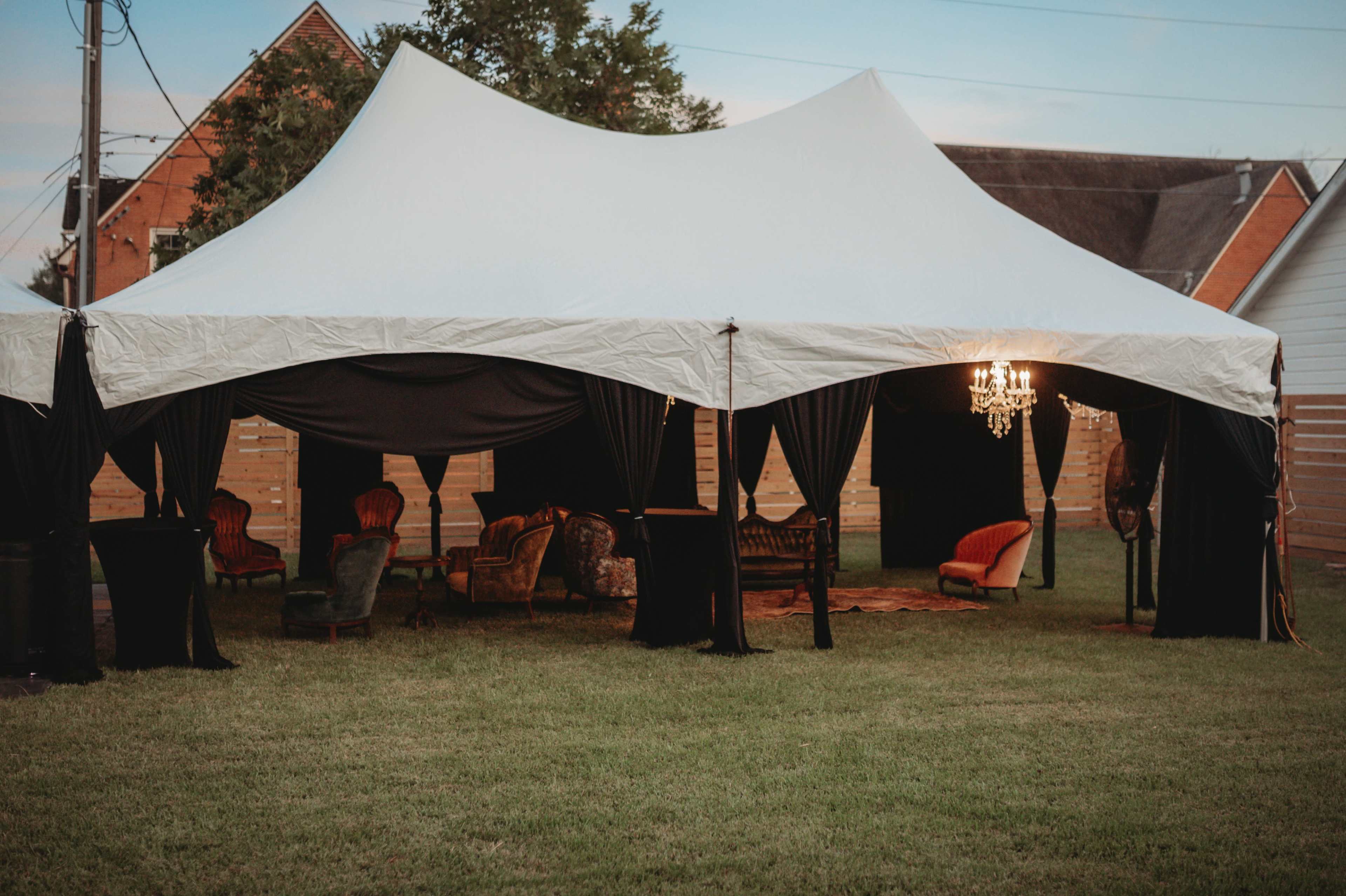 A large white tent with black drapes houses various vintage-style seating arrangements and a chandelier in an outdoor setting.