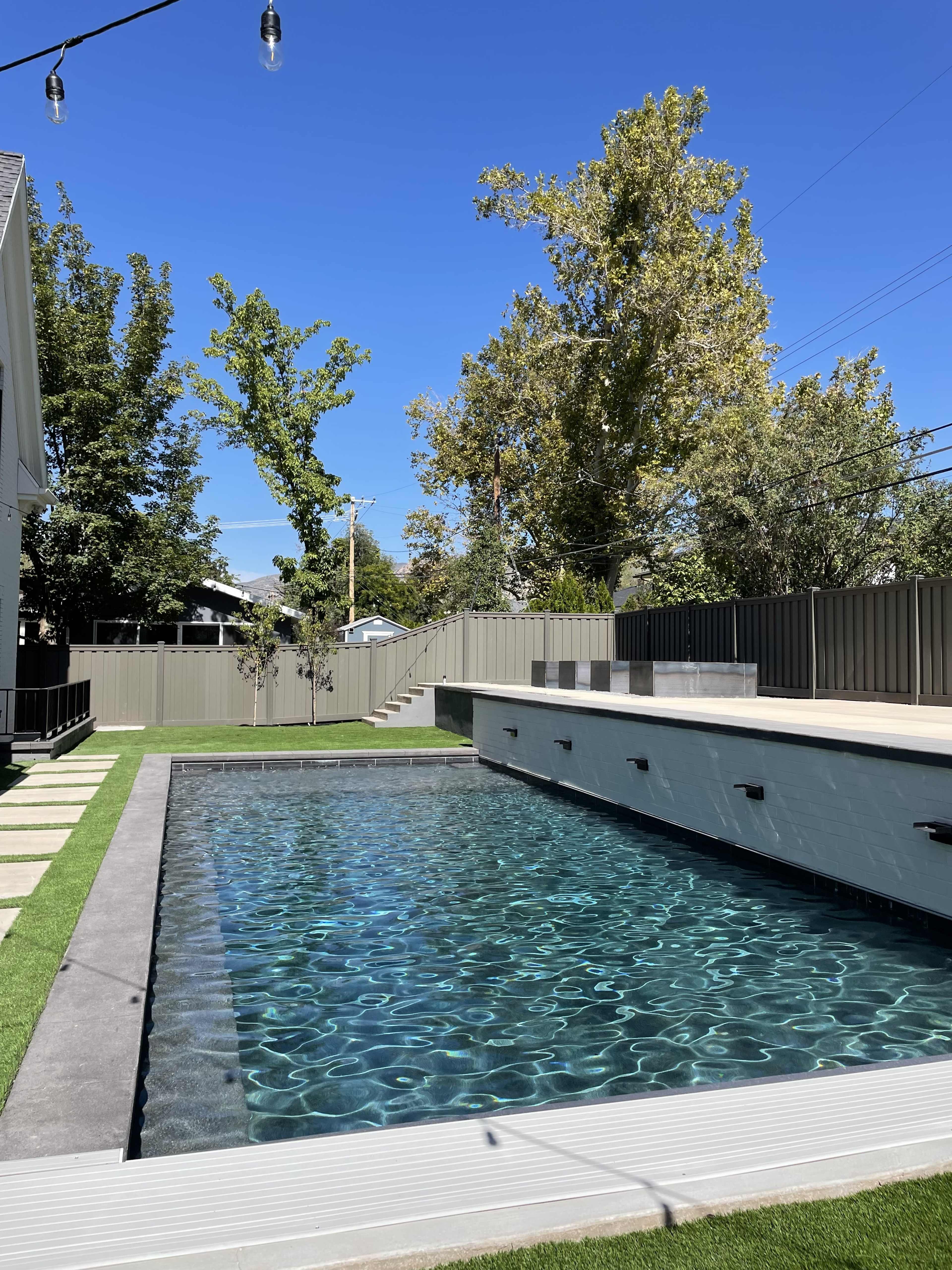 A modern swimming pool surrounded by a landscaped yard with trees and a gray privacy fence under a clear blue sky.