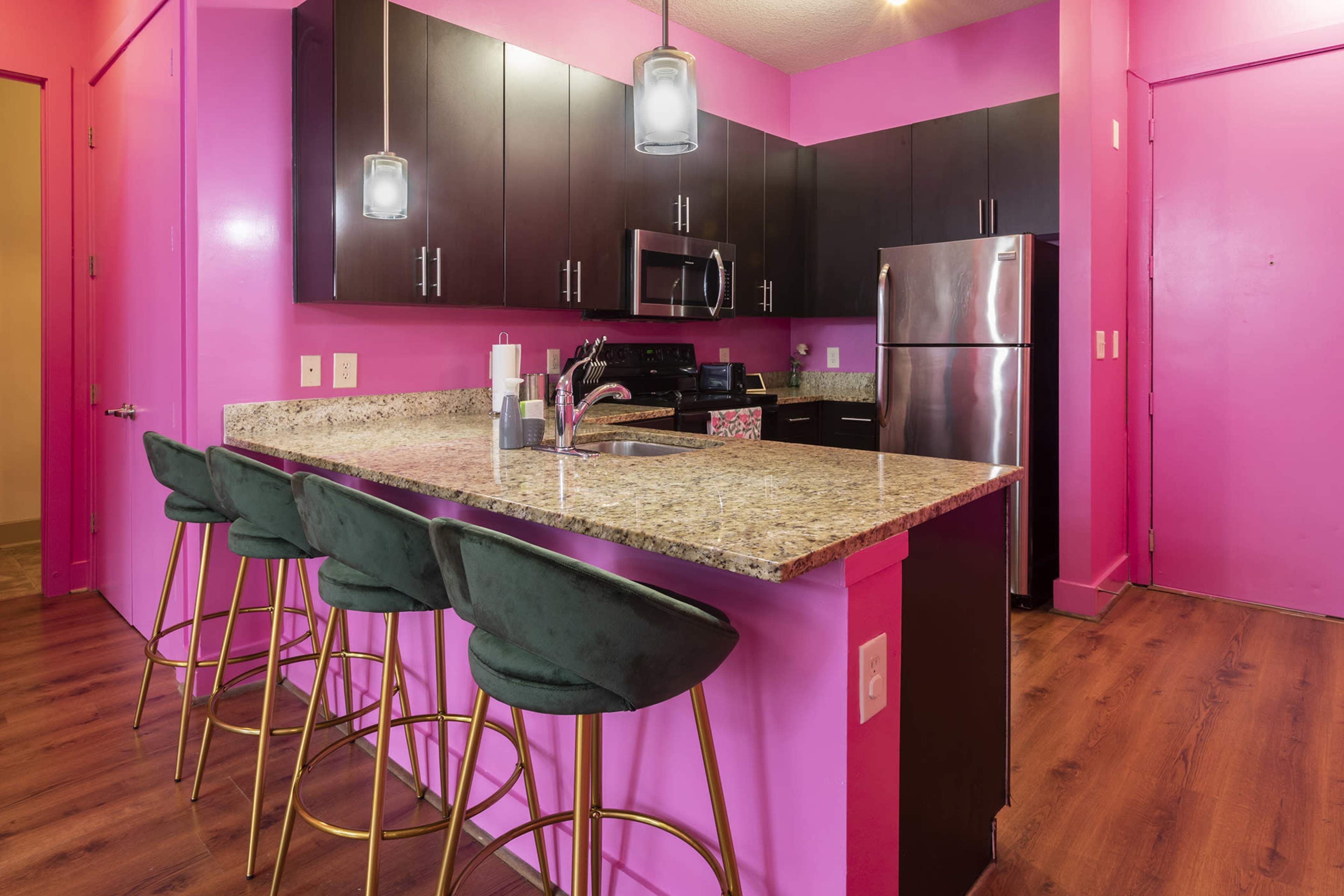 A contemporary kitchen featuring pink walls, dark cabinetry, stainless steel appliances, and a bar with four green stools.