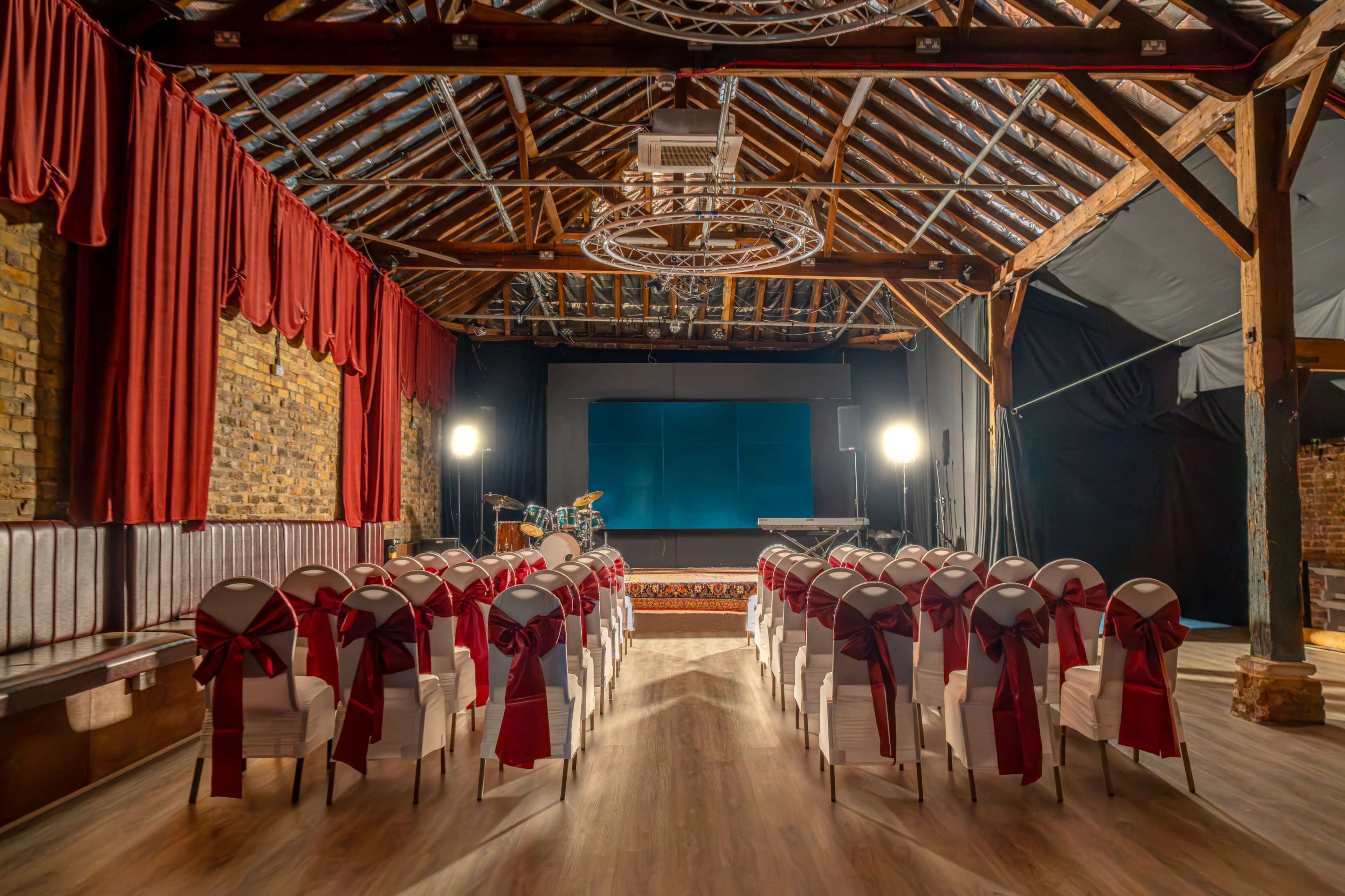 A spacious venue with wooden beams features rows of white chairs adorned with red bows, facing a stage with lighting and a band setup.