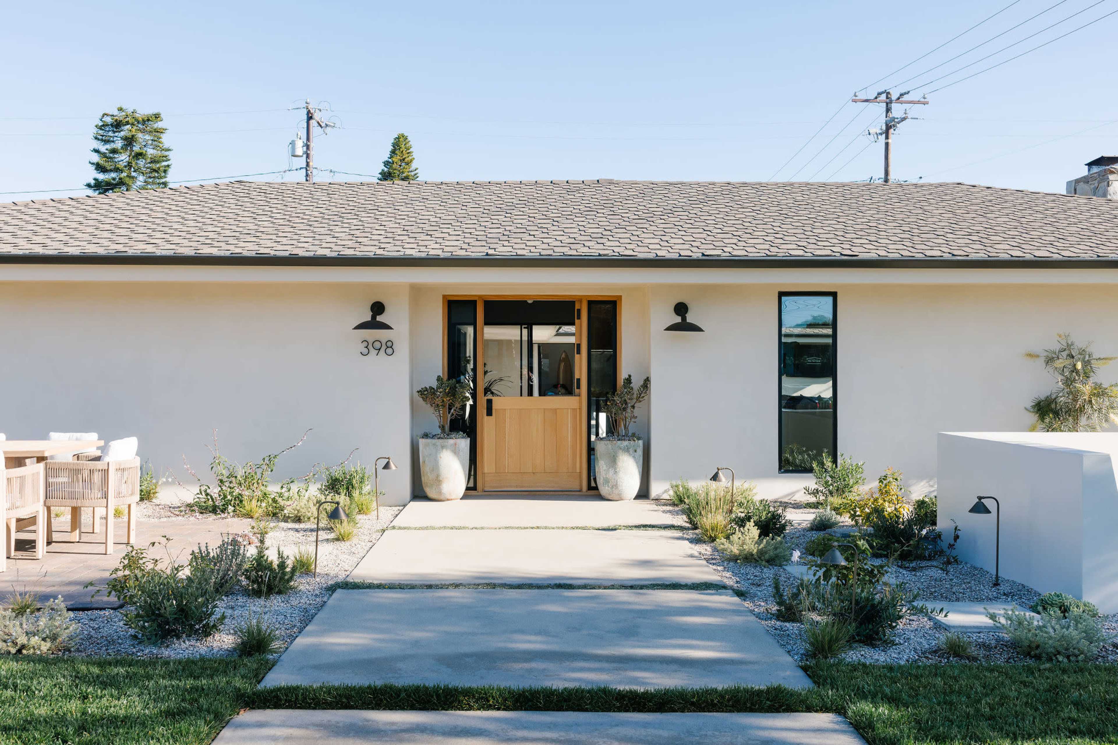 The image shows a modern single-story house with a wooden front door, landscaped pathways, and outdoor seating.