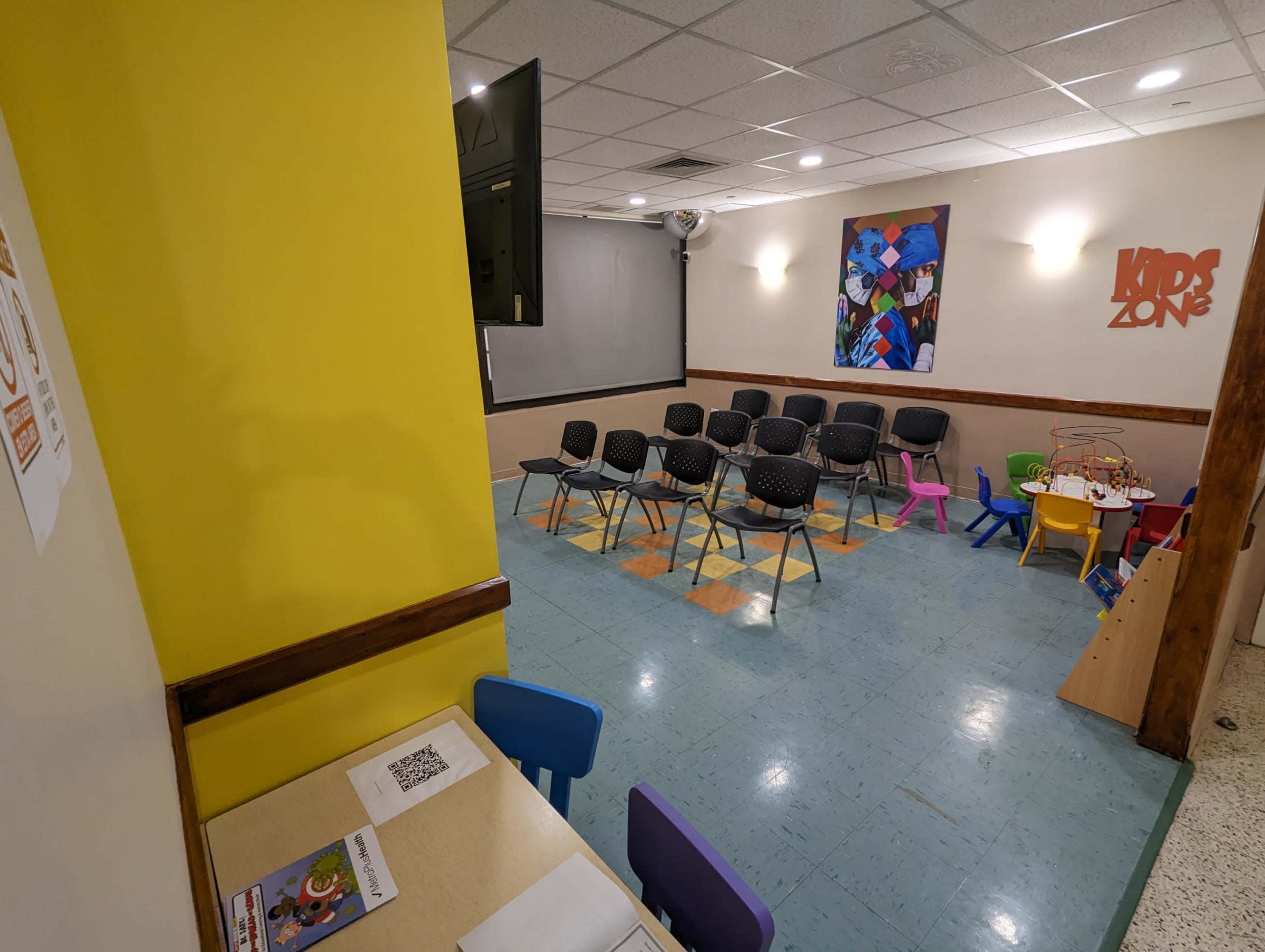 A brightly colored children's activity room features chairs arranged in rows, a small table with chairs, and a wall adorned with playful artwork.