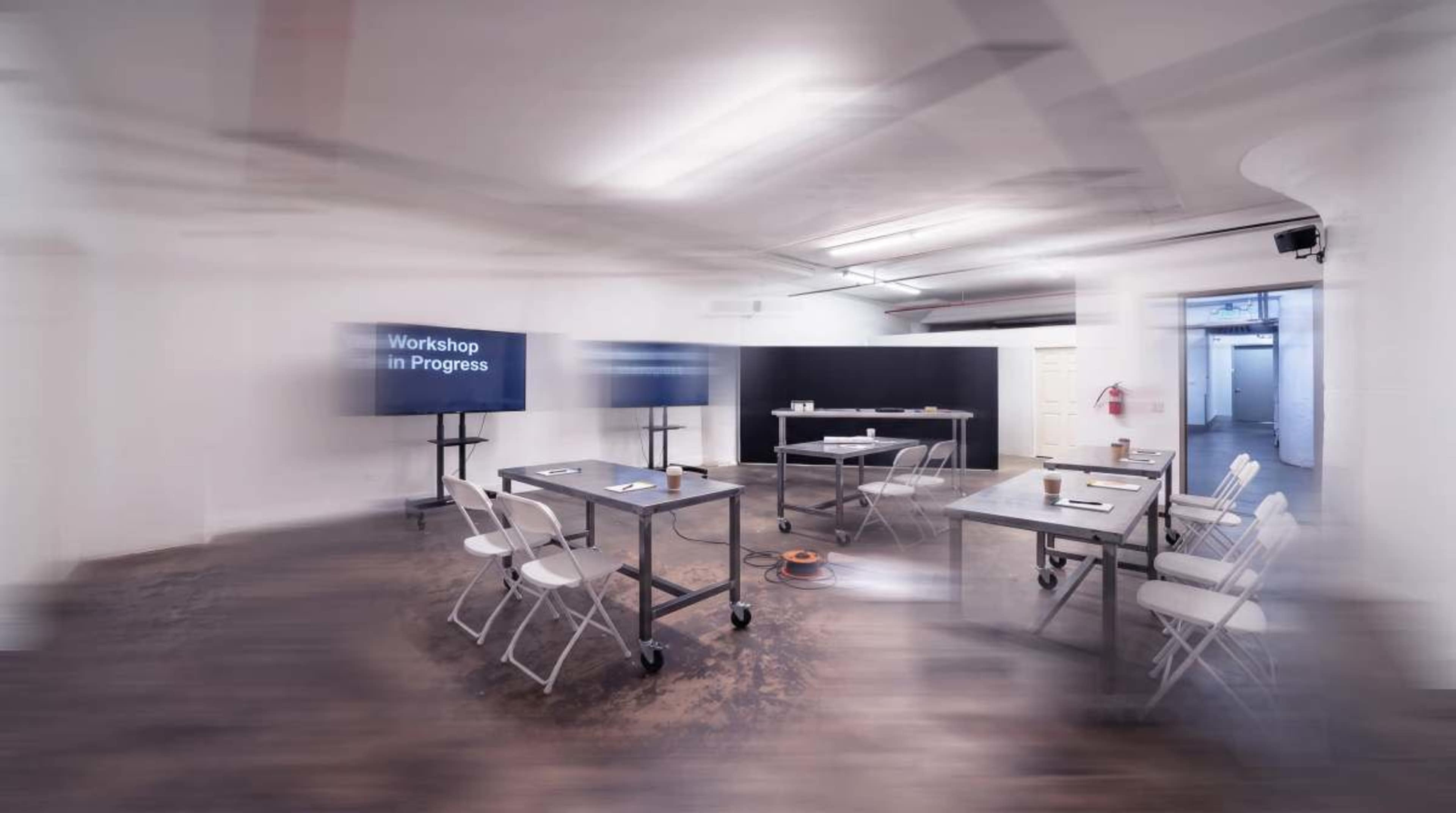 A minimalistic workshop space features several metal tables and chairs arranged around a central area, with two screens displaying "Workshop in Progress" on the walls.
