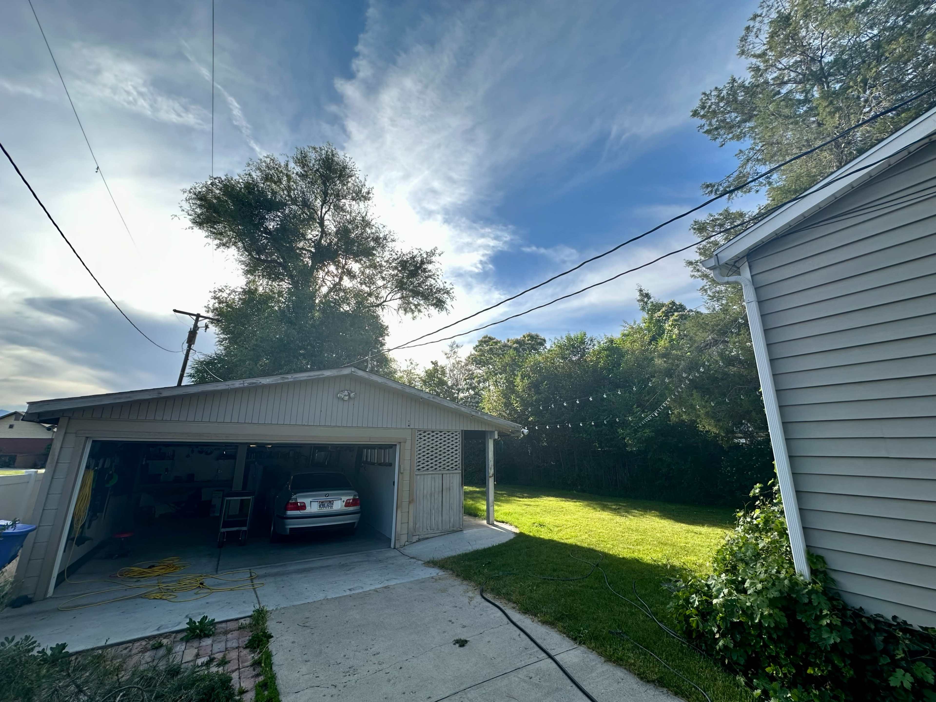 A garage with a parked car is situated next to a grassy area and surrounded by trees under a partly cloudy sky.