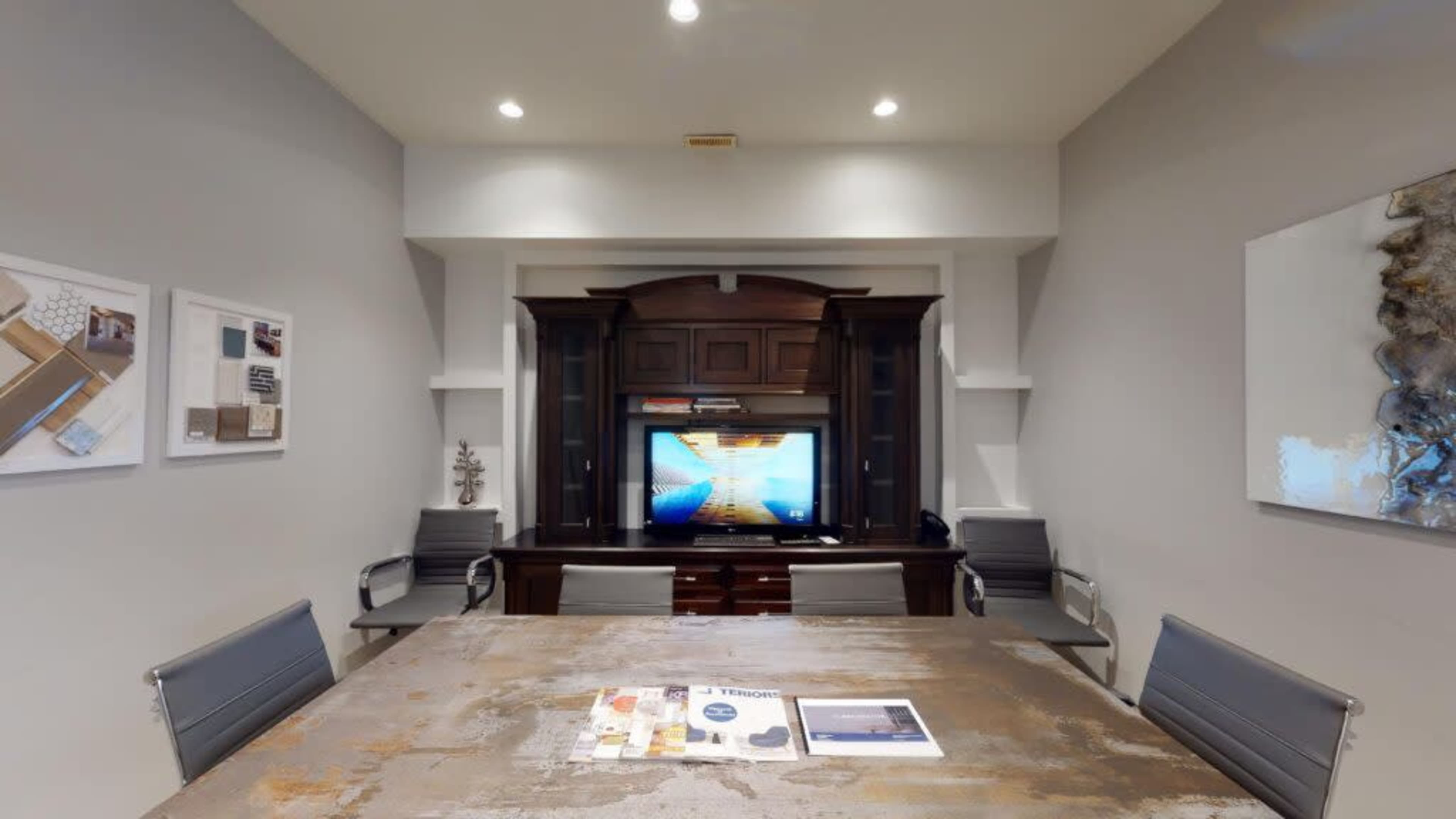 The image shows a modern conference room with a large wooden table, metal chairs, and a wall-mounted television displaying a graphic on a media console.