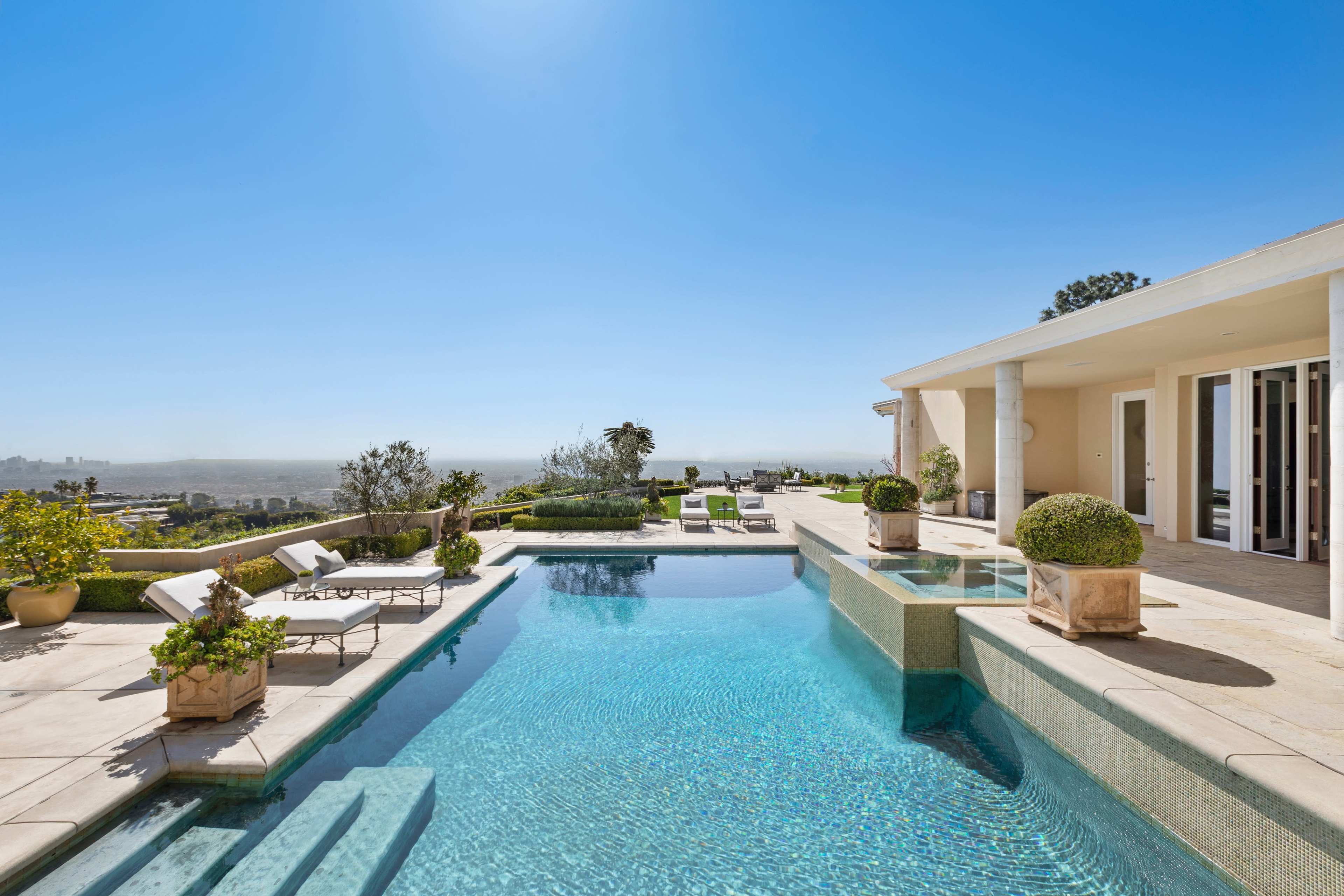 A modern swimming pool with a spa area is set in a landscaped backyard overlooking a panoramic city view.