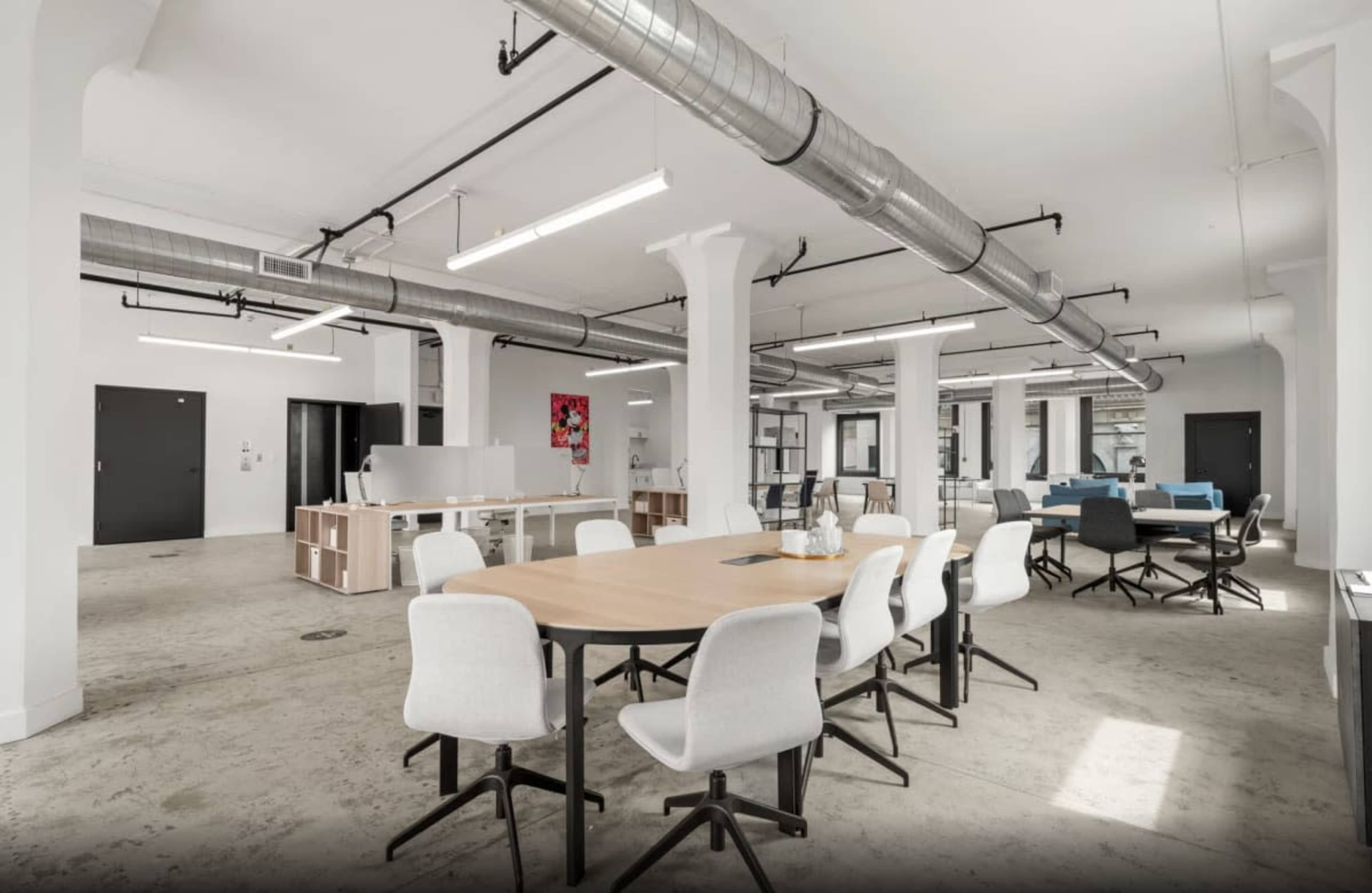 The image shows a spacious, modern office environment with multiple tables and chairs arranged for collaboration, featuring exposed ductwork and large windows.