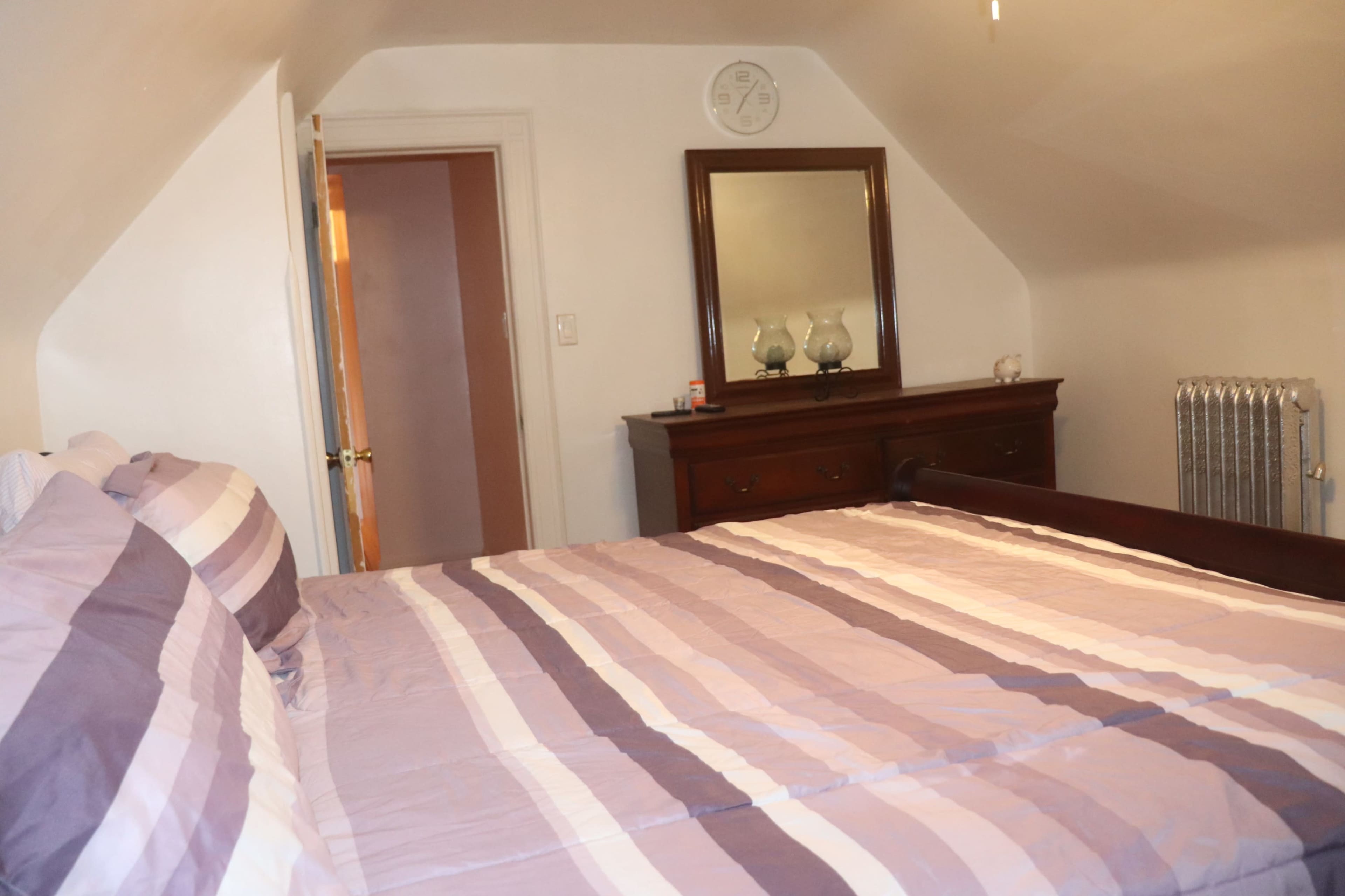 A bedroom features a bed with striped linens and a wooden dresser against the wall under a sloped ceiling.