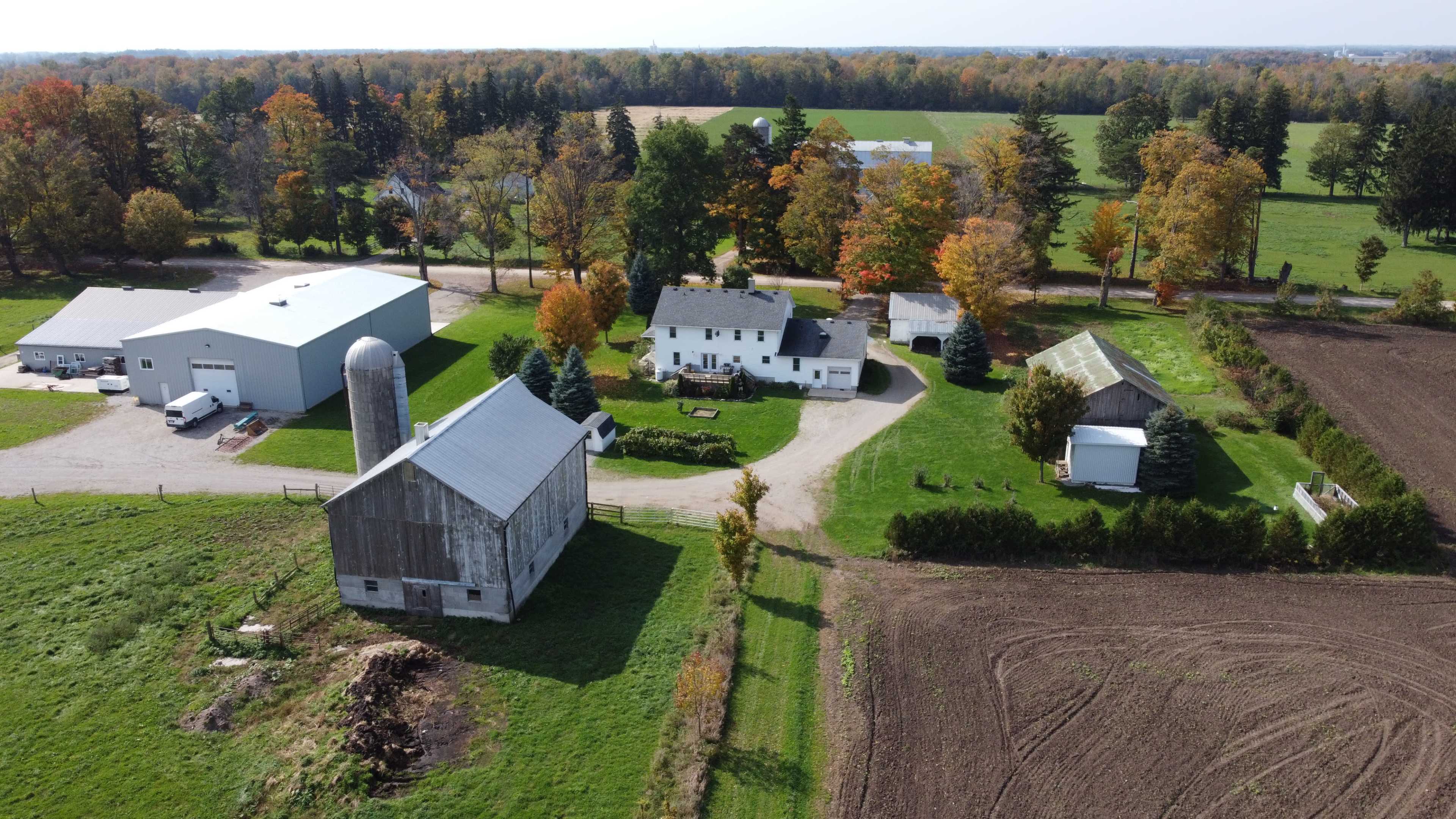 An aerial view of a farm featuring multiple buildings, including a barn, silos, and a white house, surrounded by fields and trees with autumn foliage.