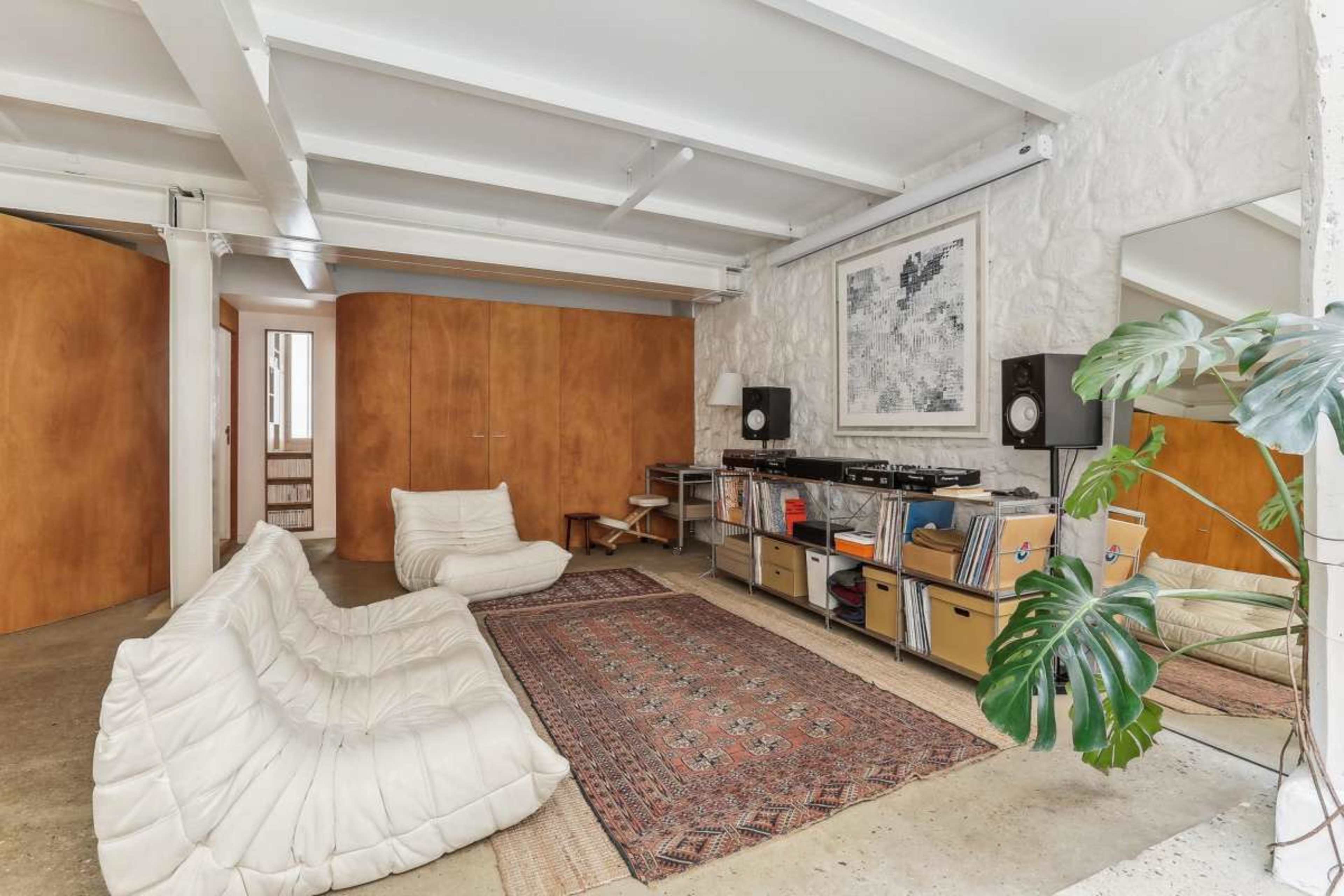 The image shows a spacious living room with a white, cushioned sofa set, a wooden storage unit, and a record player setup against a textured stone wall.
