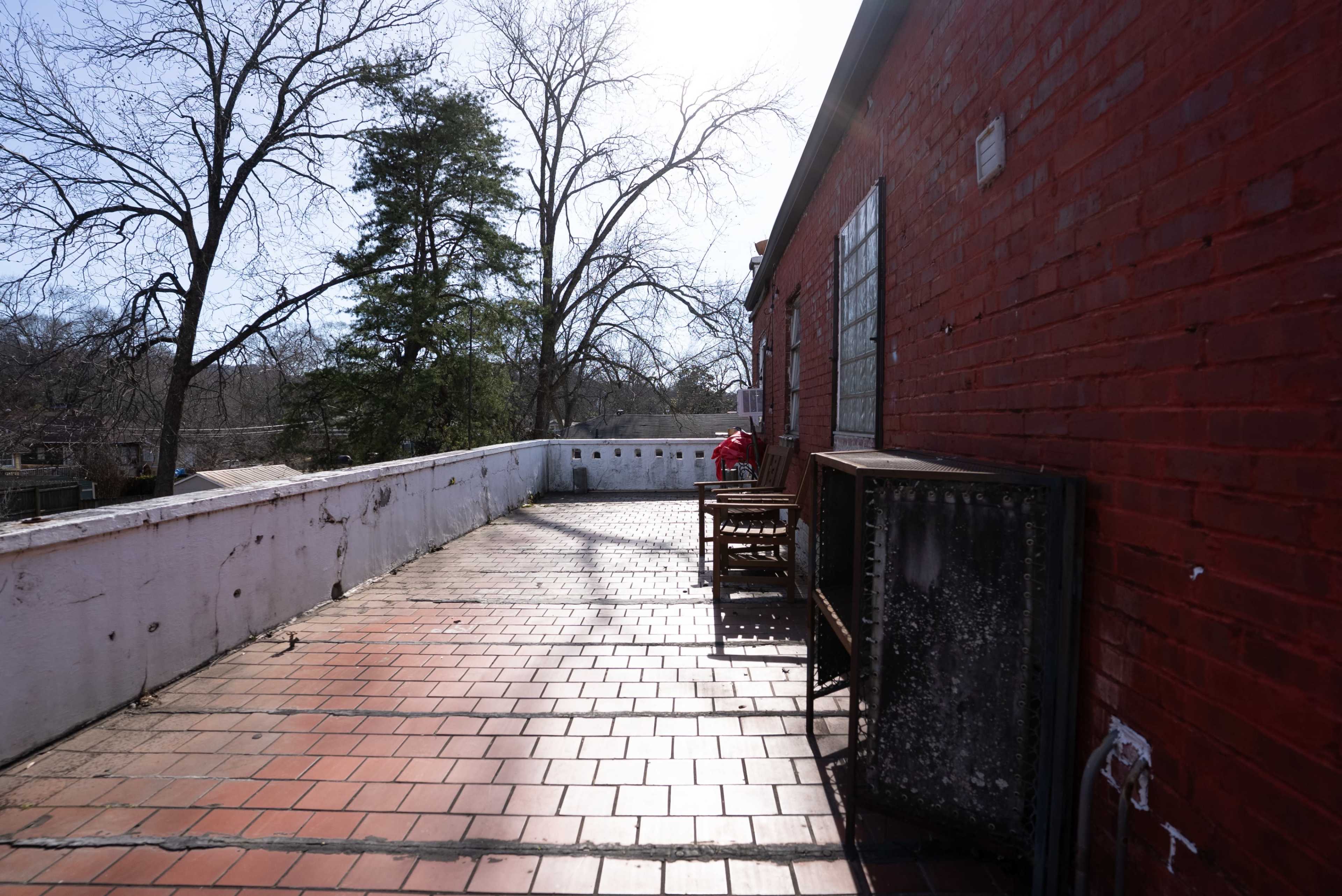 A sunlit terrace features a brick wall and wooden chairs, surrounded by bare trees and a clear sky.
