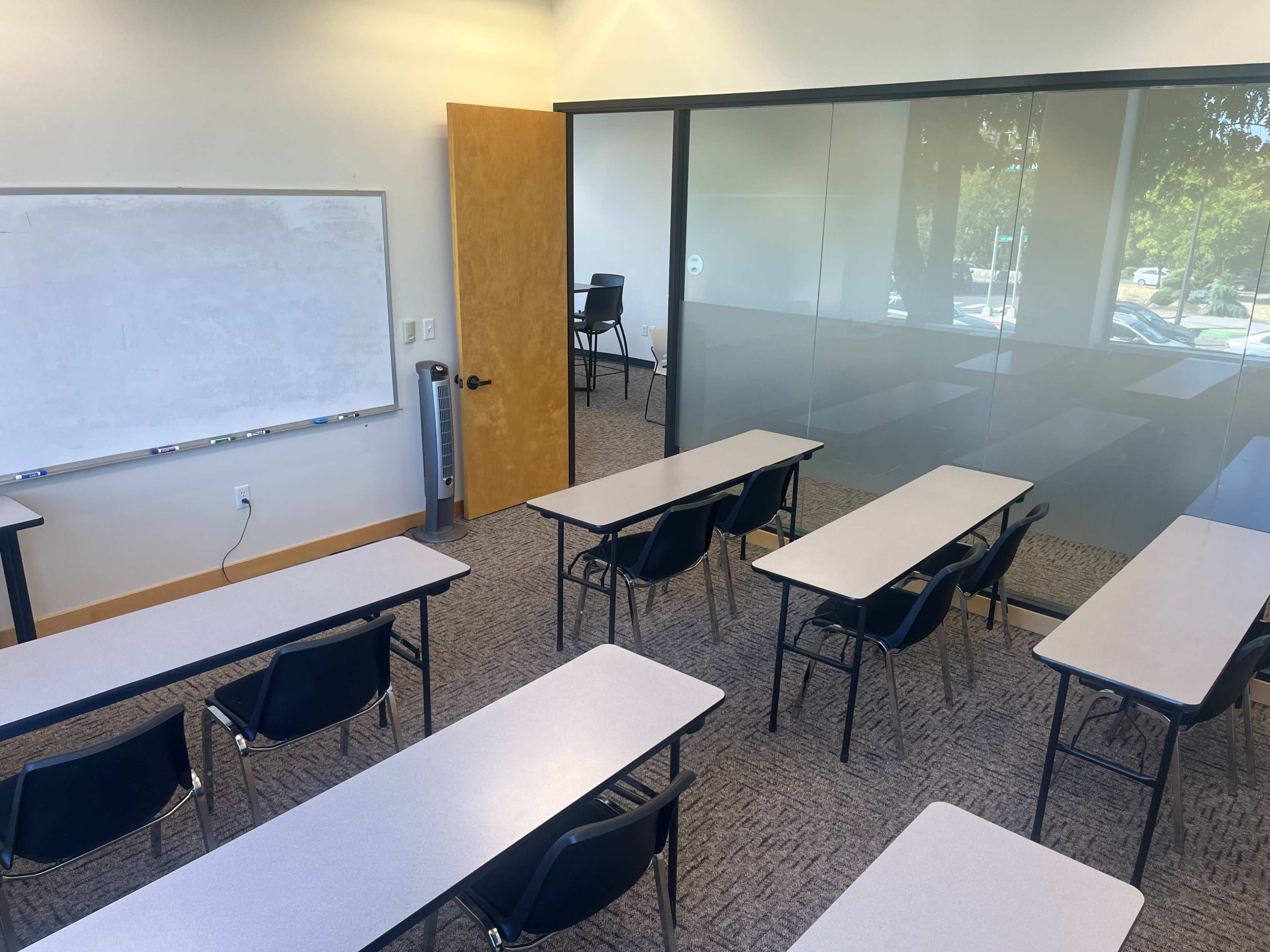 The image shows a classroom with several rows of desks facing a whiteboard, and a glass-walled section in the background containing a single chair.