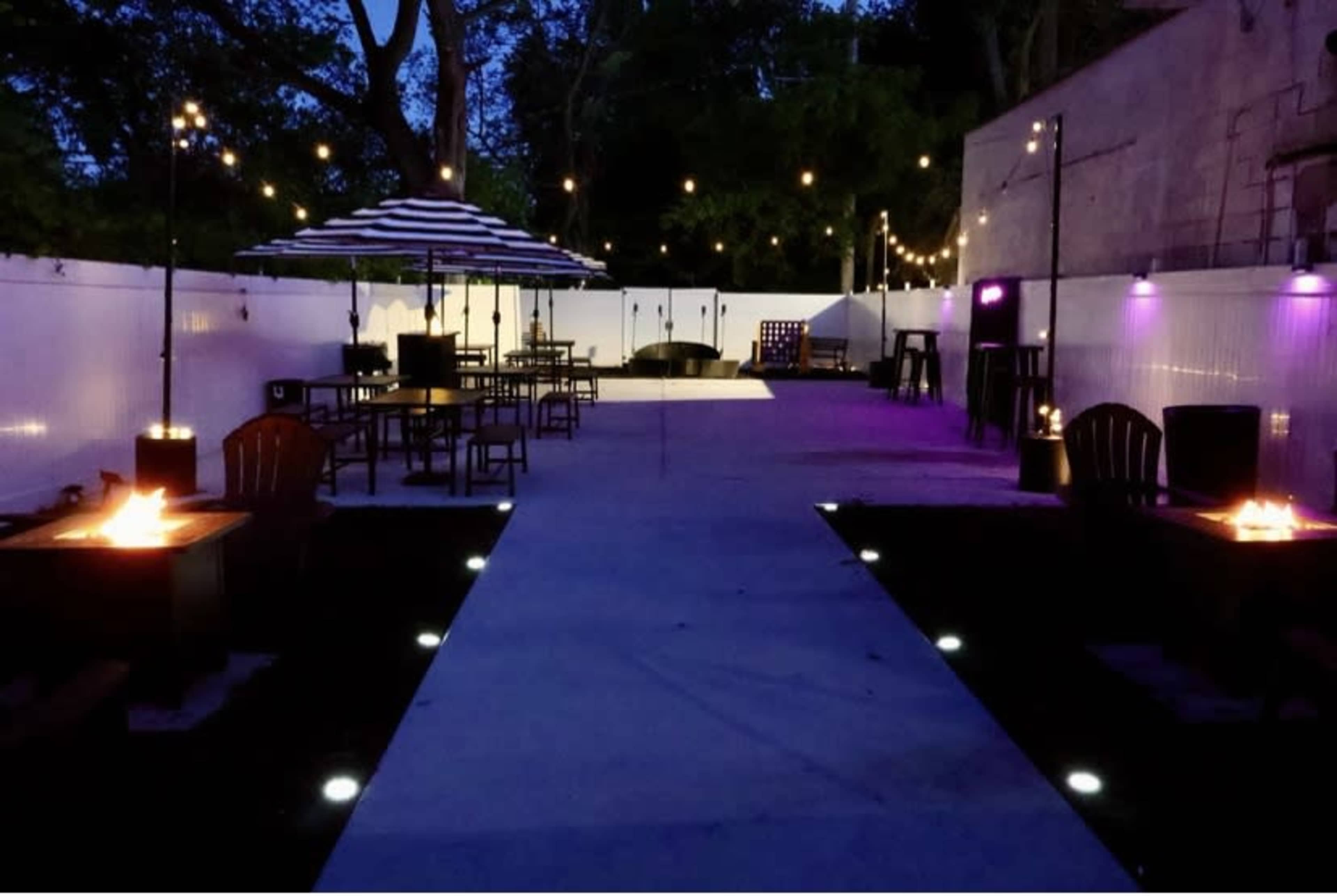 The image shows an outdoor patio area illuminated by string lights, with fire pits and tables arranged along a concrete pathway.