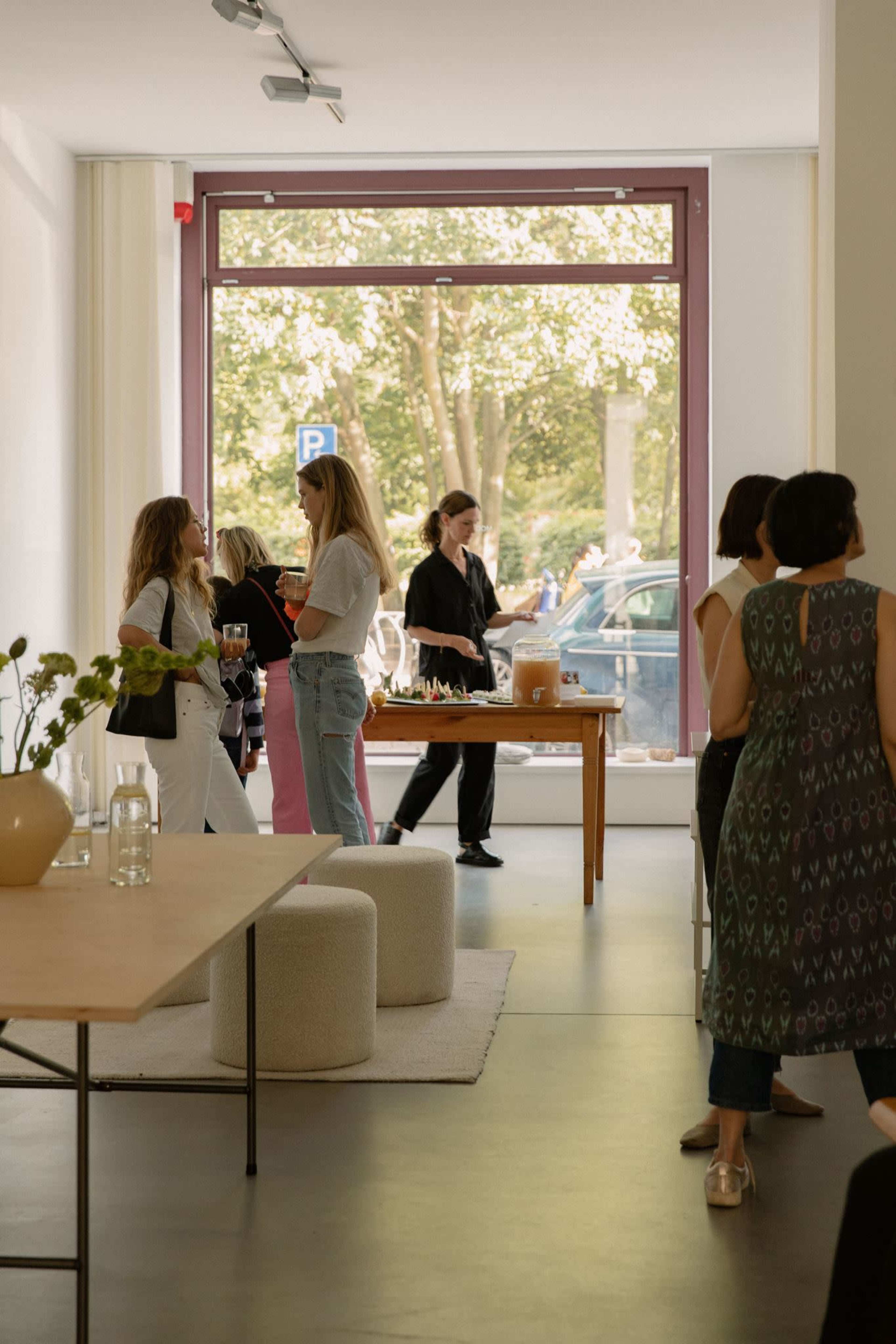 A group of people socialize in a bright, modern interior with large windows and a table set for refreshments.