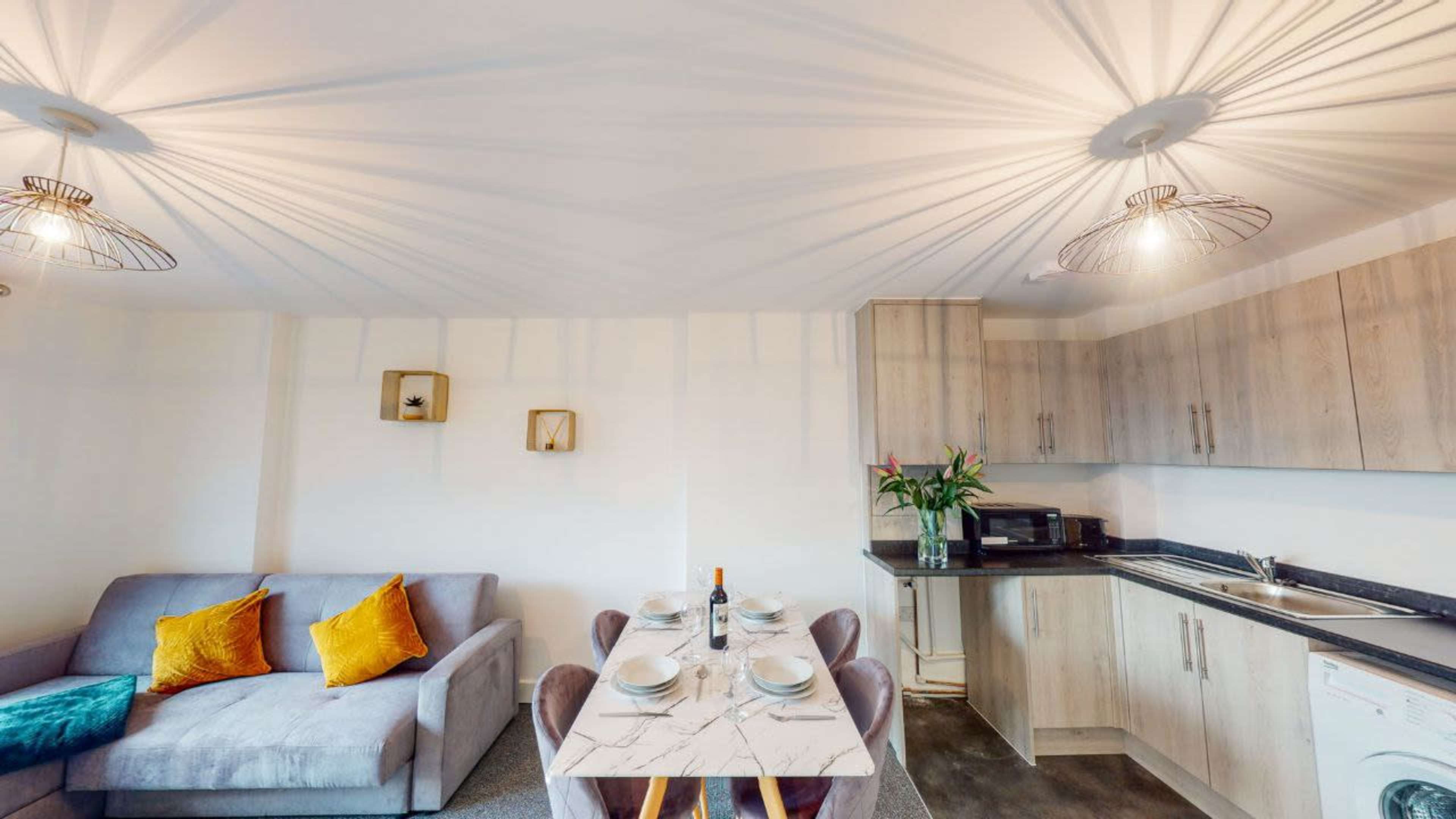 A modern kitchen and dining area feature a marble table set for four, a gray sofa with colorful cushions, and pendant lighting casting shadows on the ceiling.