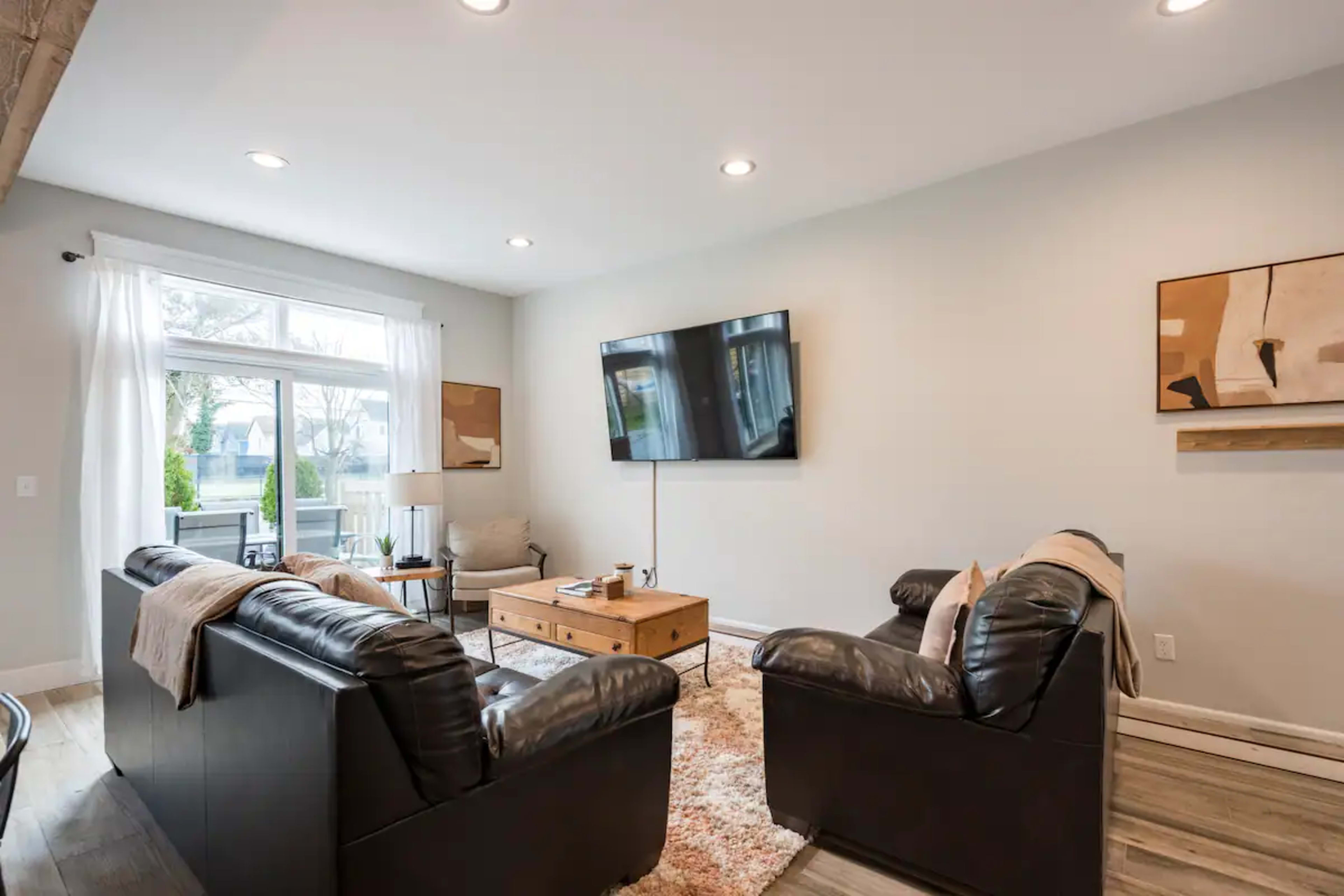 The living room features two black leather sofas facing a coffee table, with a large flat-screen TV mounted on the wall and windows allowing natural light to enter.