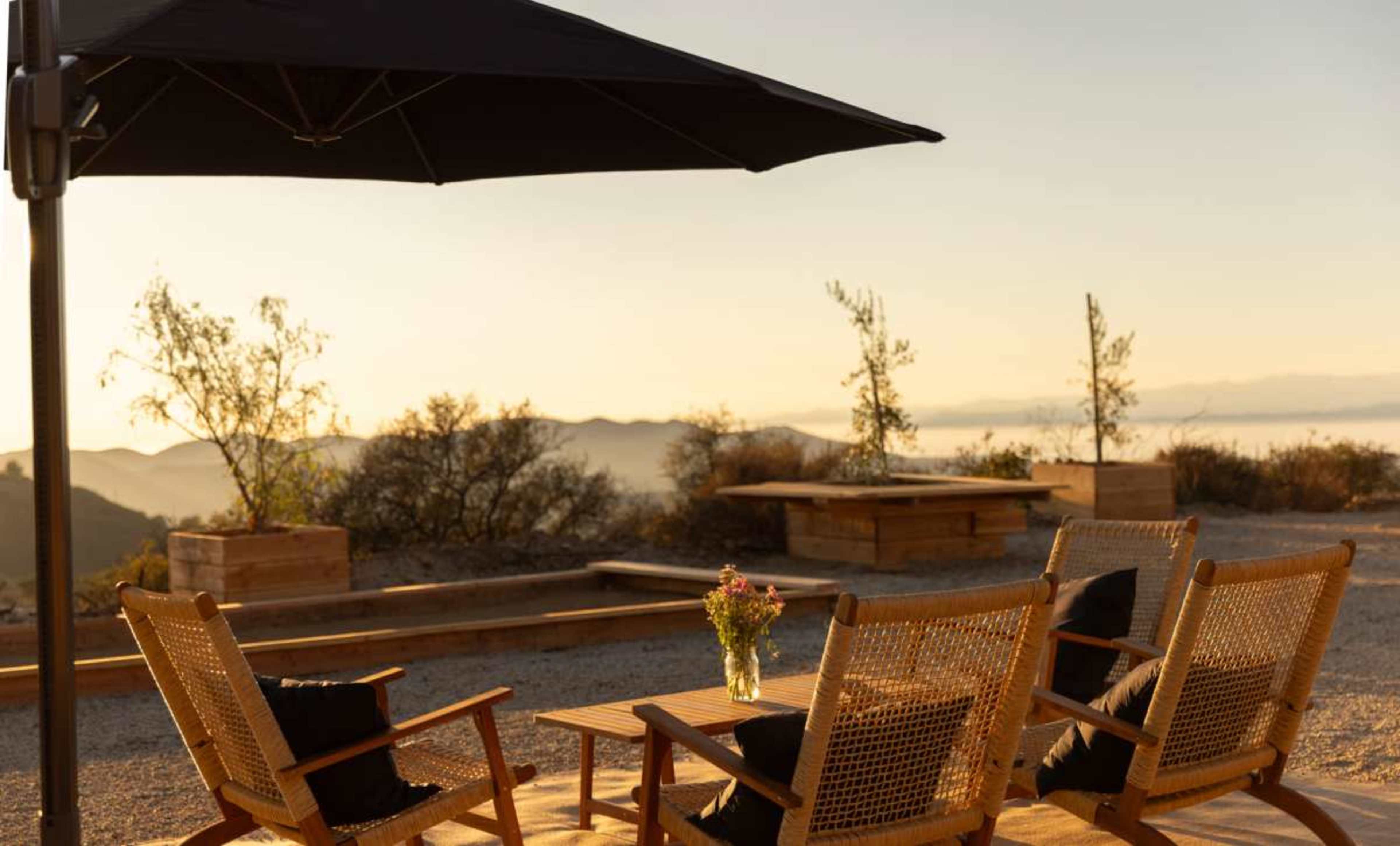 Three wooden chairs with black cushions are arranged around a small table under a large umbrella, set against a backdrop of hills at sunset.