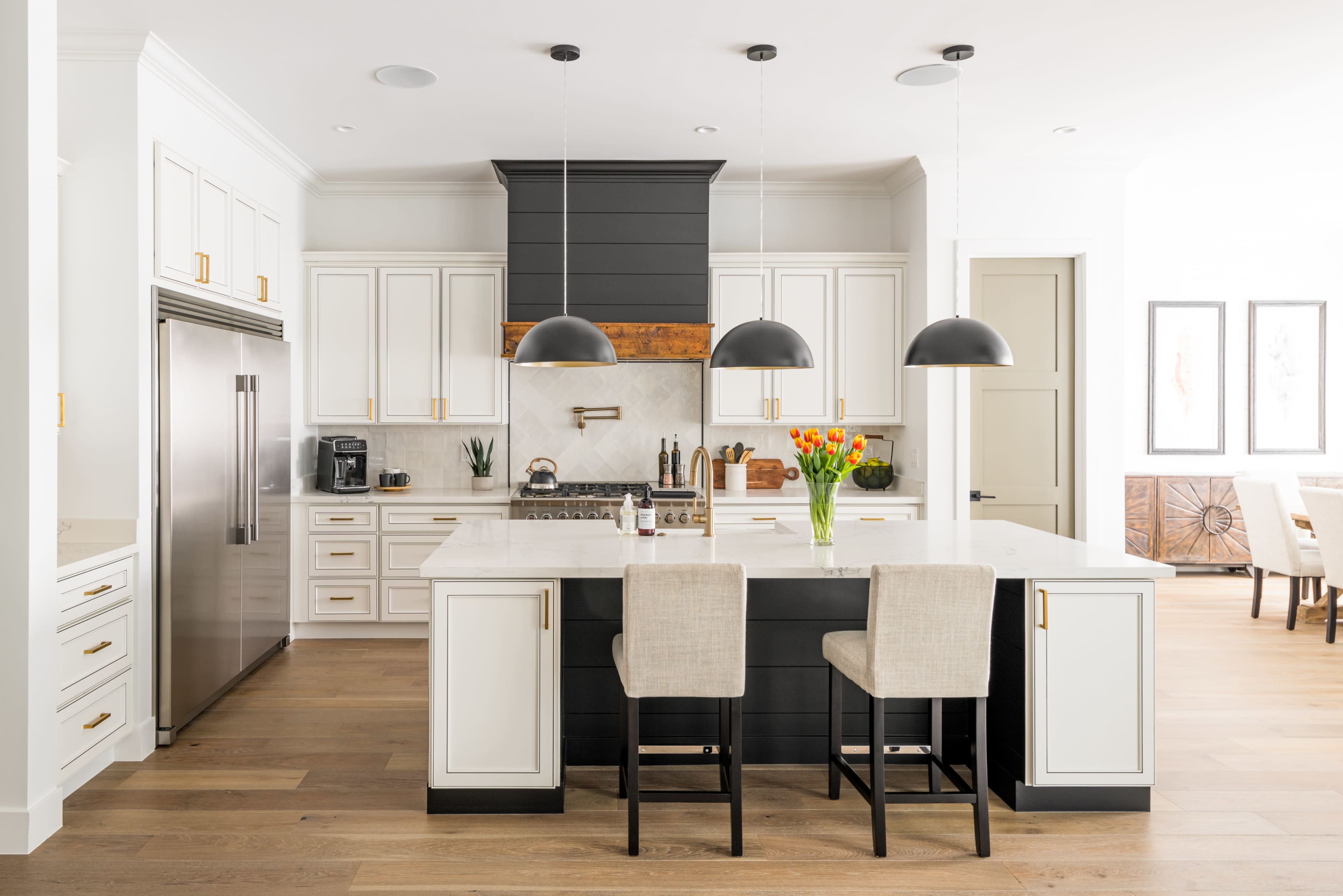 A modern kitchen features white cabinetry, a dark island with two bar stools, granite countertops, and black pendant lights.
