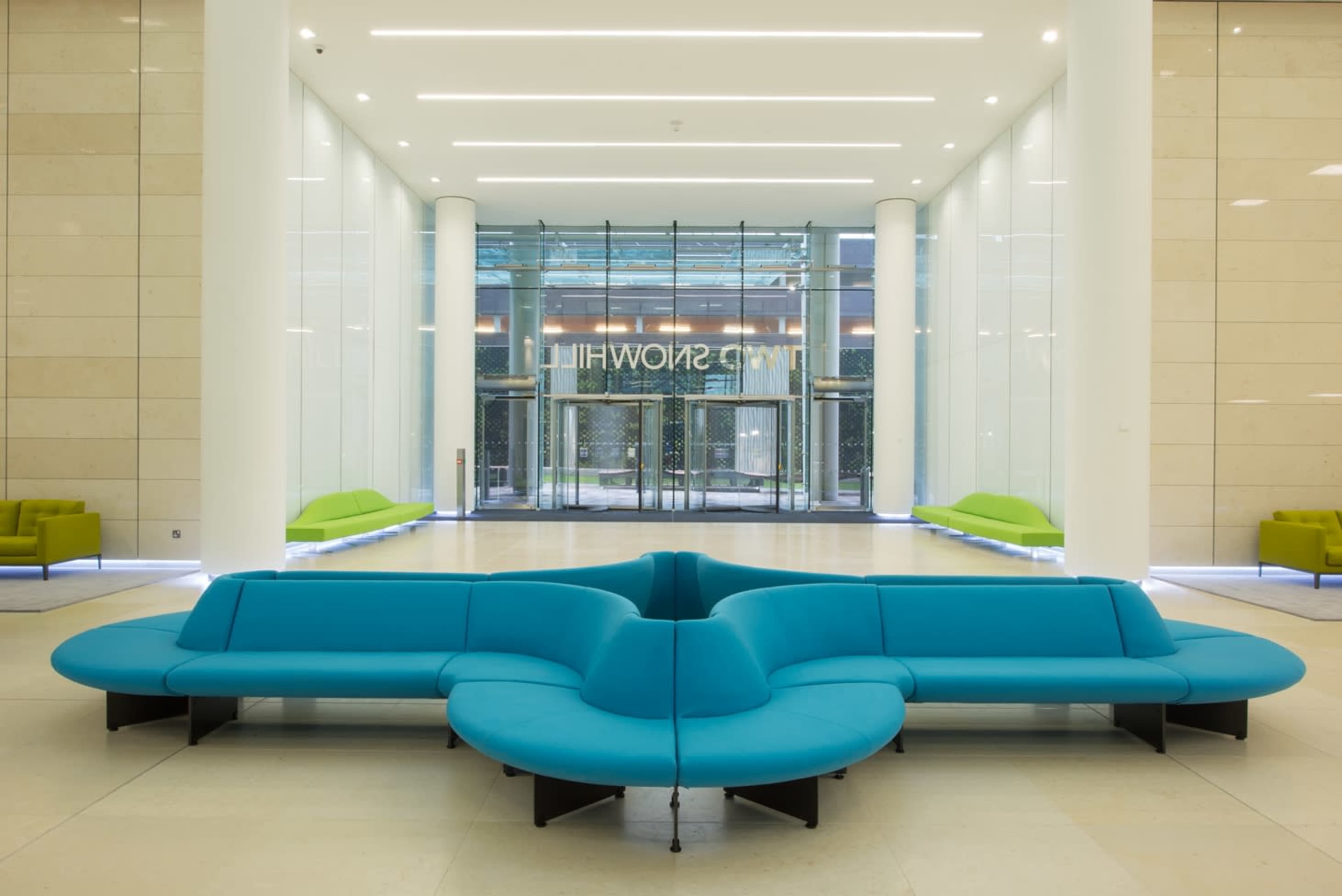 A modern lobby with a large turquoise sofa arranged in an abstract shape, flanked by green chairs, and glass doors leading outside.