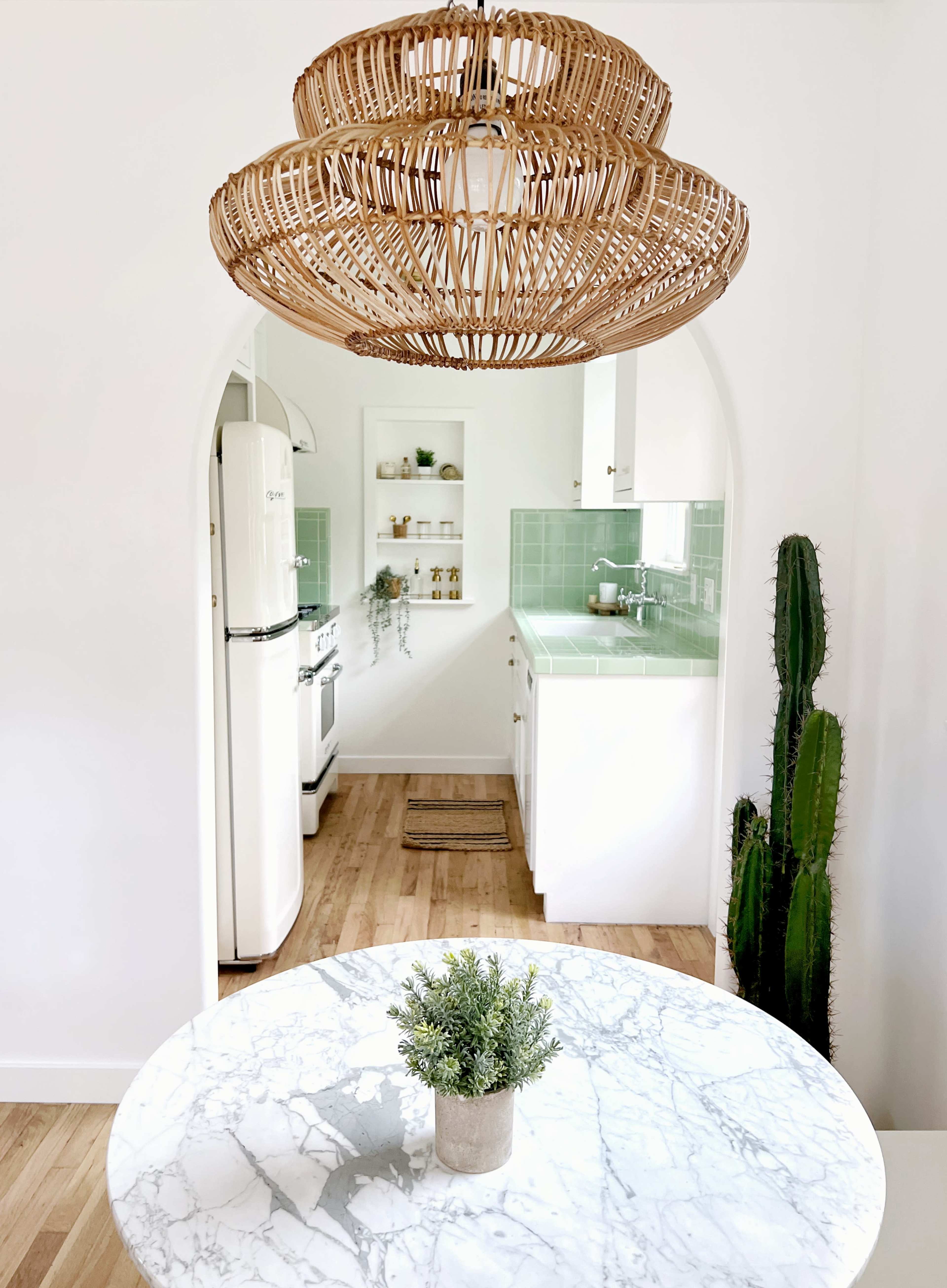 A kitchen with a marble table, a large woven pendant light, and green tile accents is viewed through an arched entryway, featuring a cactus on the side.