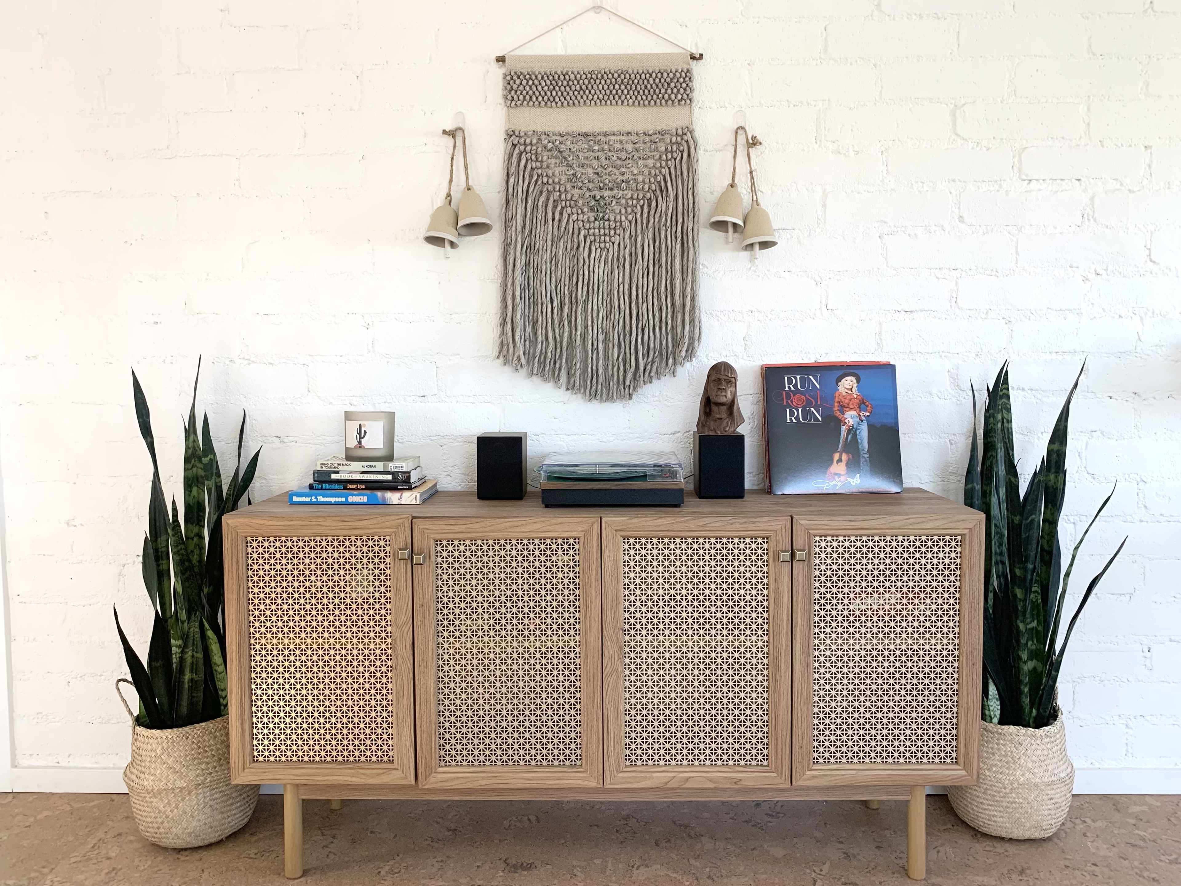 A wooden sideboard with three woven-front cabinets is placed against a white brick wall, accompanied by two tall plants in woven baskets and decorative items on top, including books and a magazine.