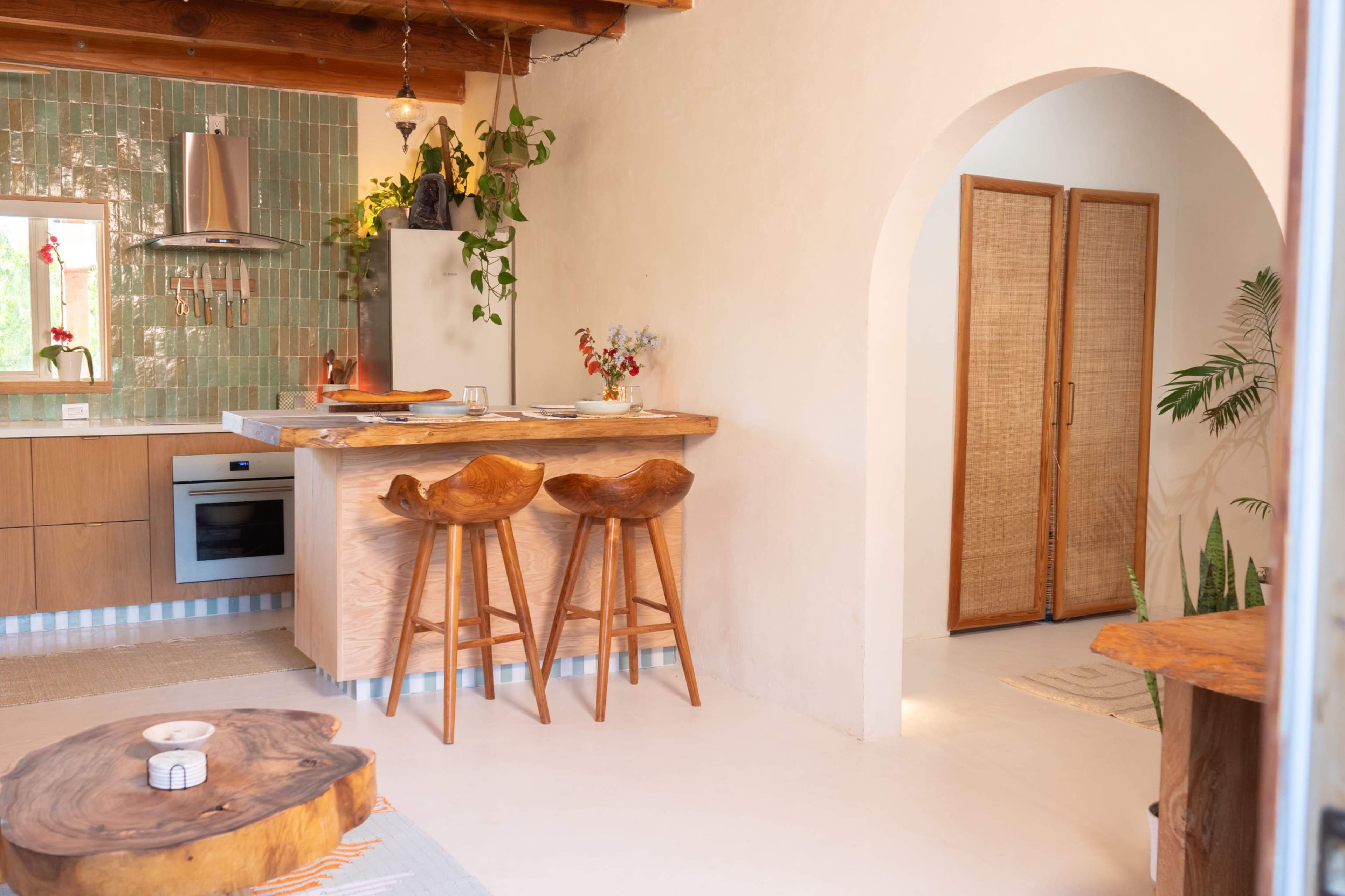 A modern kitchen features wooden stools at a counter, green tile backsplash, and natural light illuminating the space.