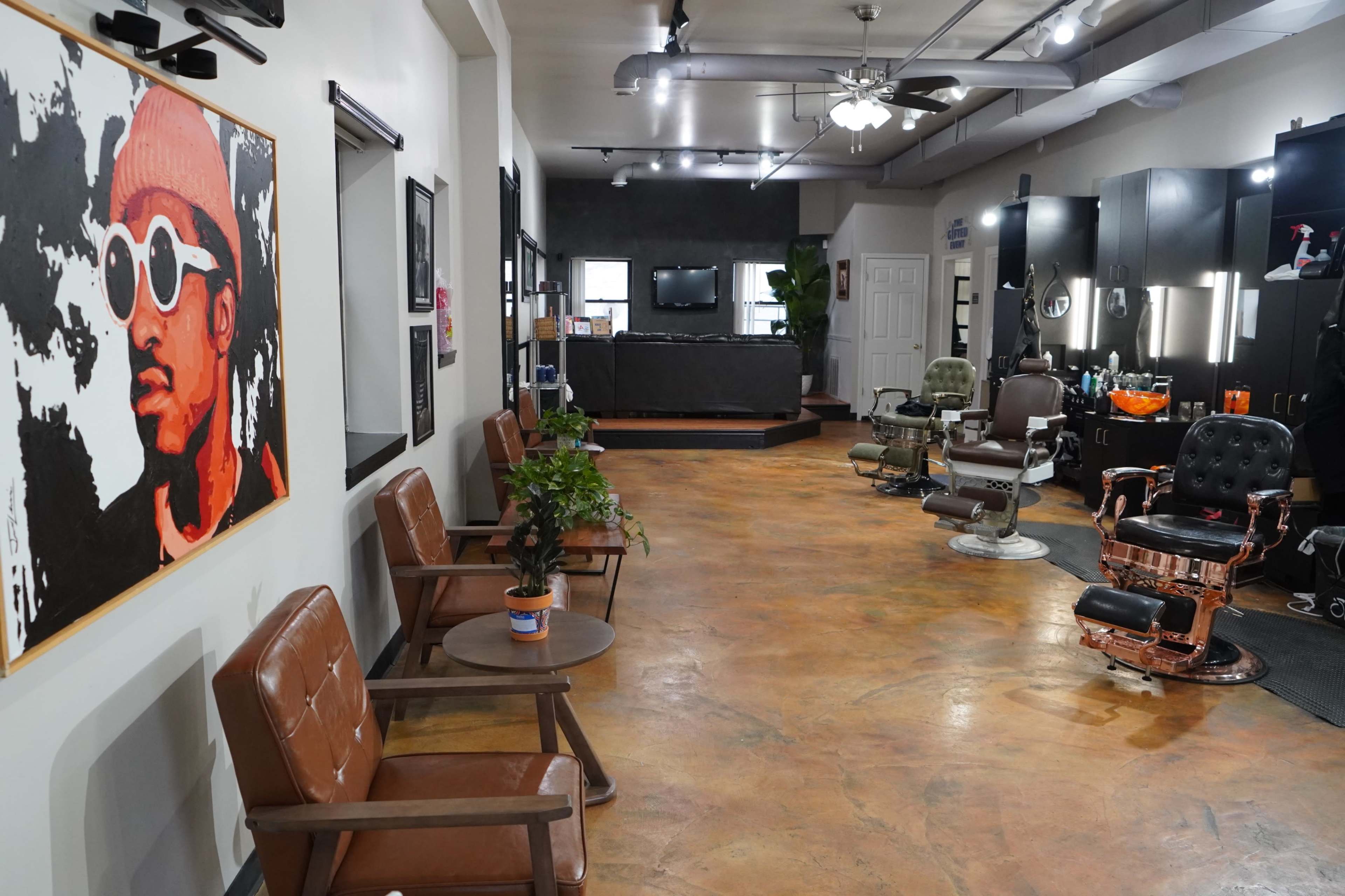 A modern barbershop interior with waiting chairs, styling stations, and a large artwork on the wall featuring a person wearing sunglasses.