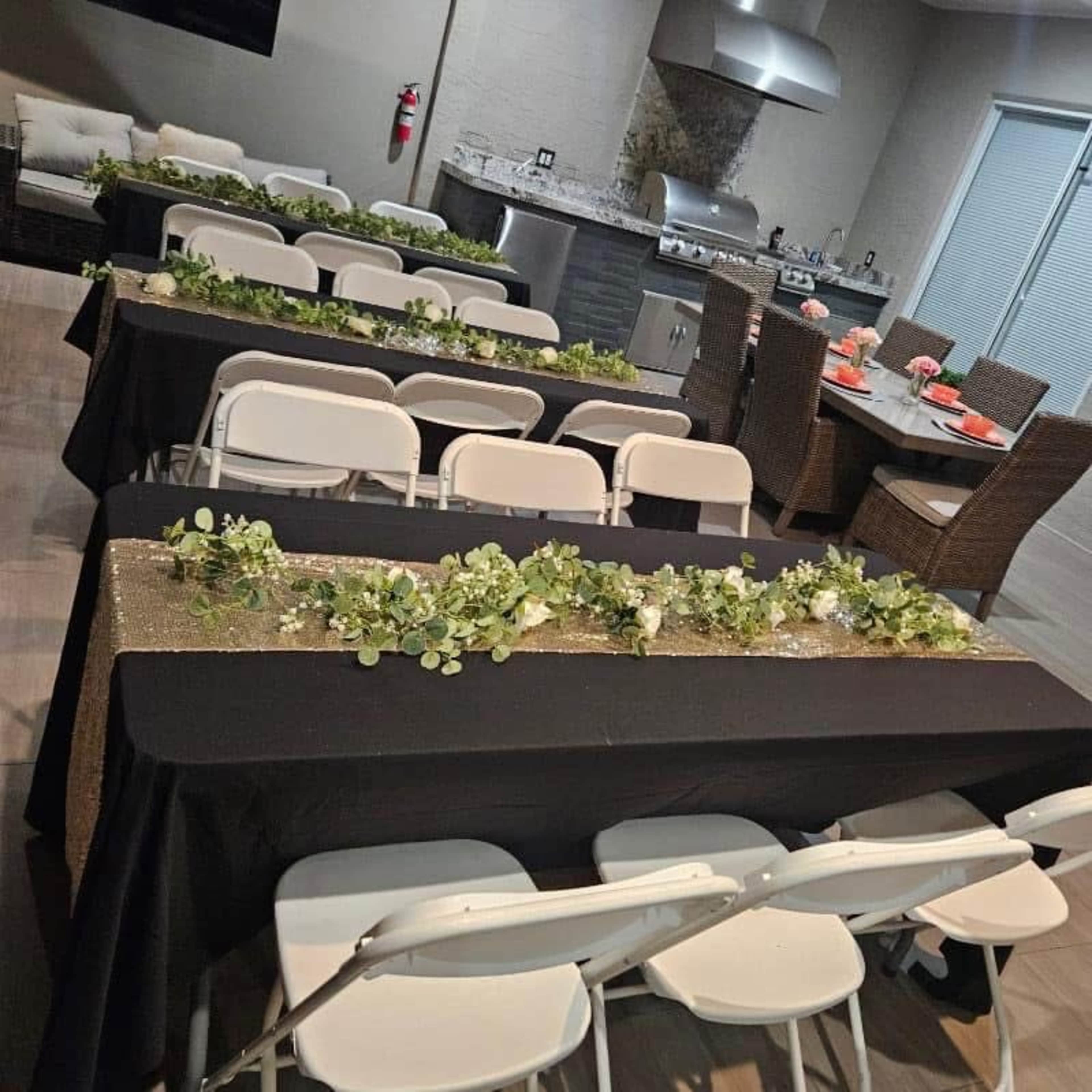 A dining setup features several long tables covered with black tablecloths adorned with greenery, arranged in a well-lit room near a kitchen area.