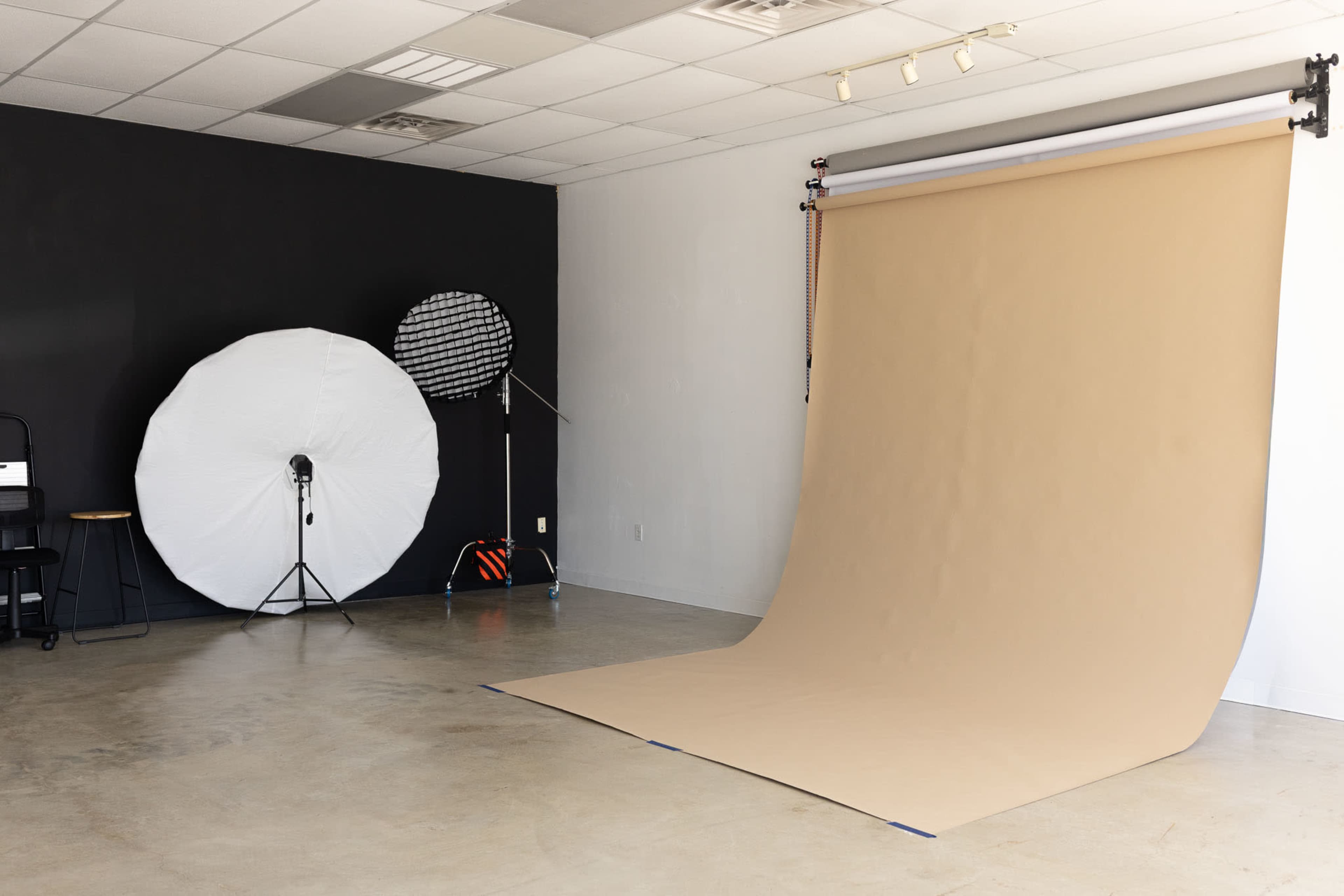 The image shows a photography studio with a large, tan seamless backdrop, a circular softbox, and a stool against a black wall.