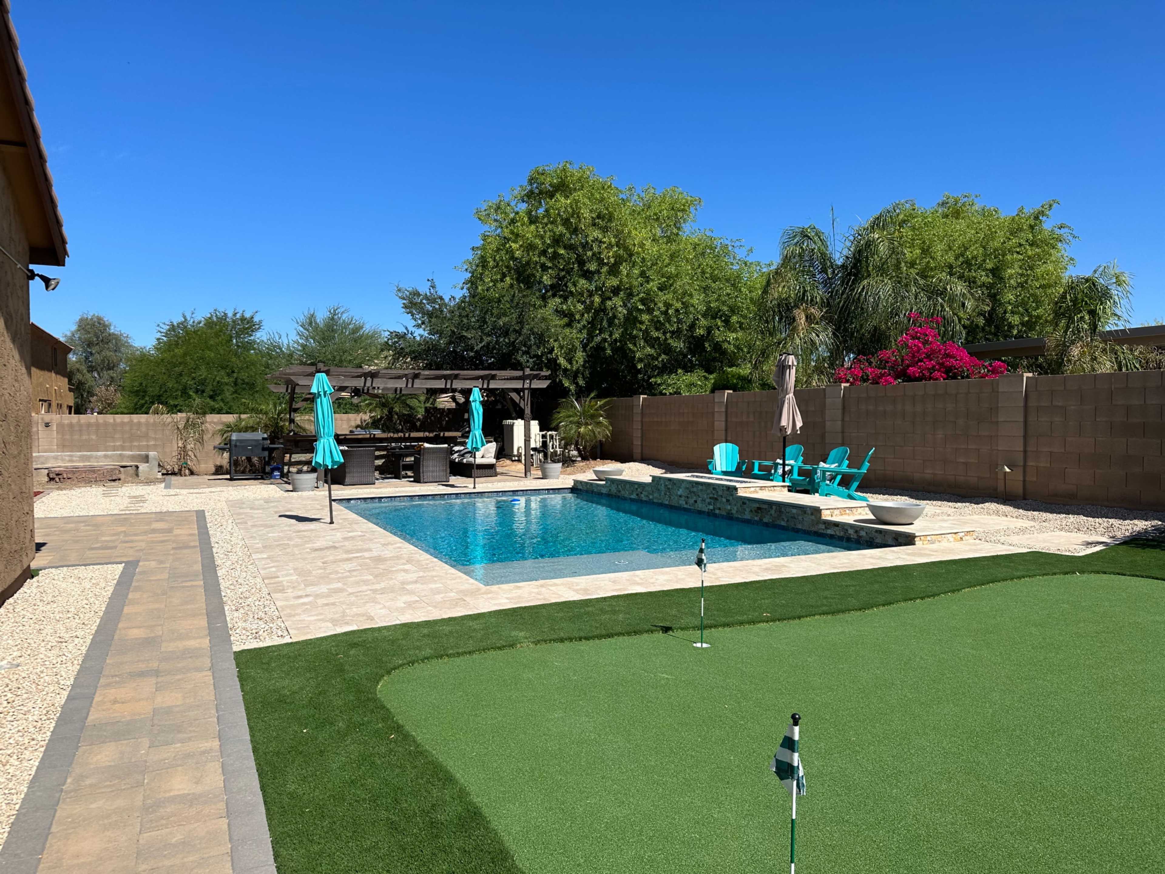 The image shows a backyard with a swimming pool, lounge chairs, a putting green, and landscaping, framed by clear blue skies.