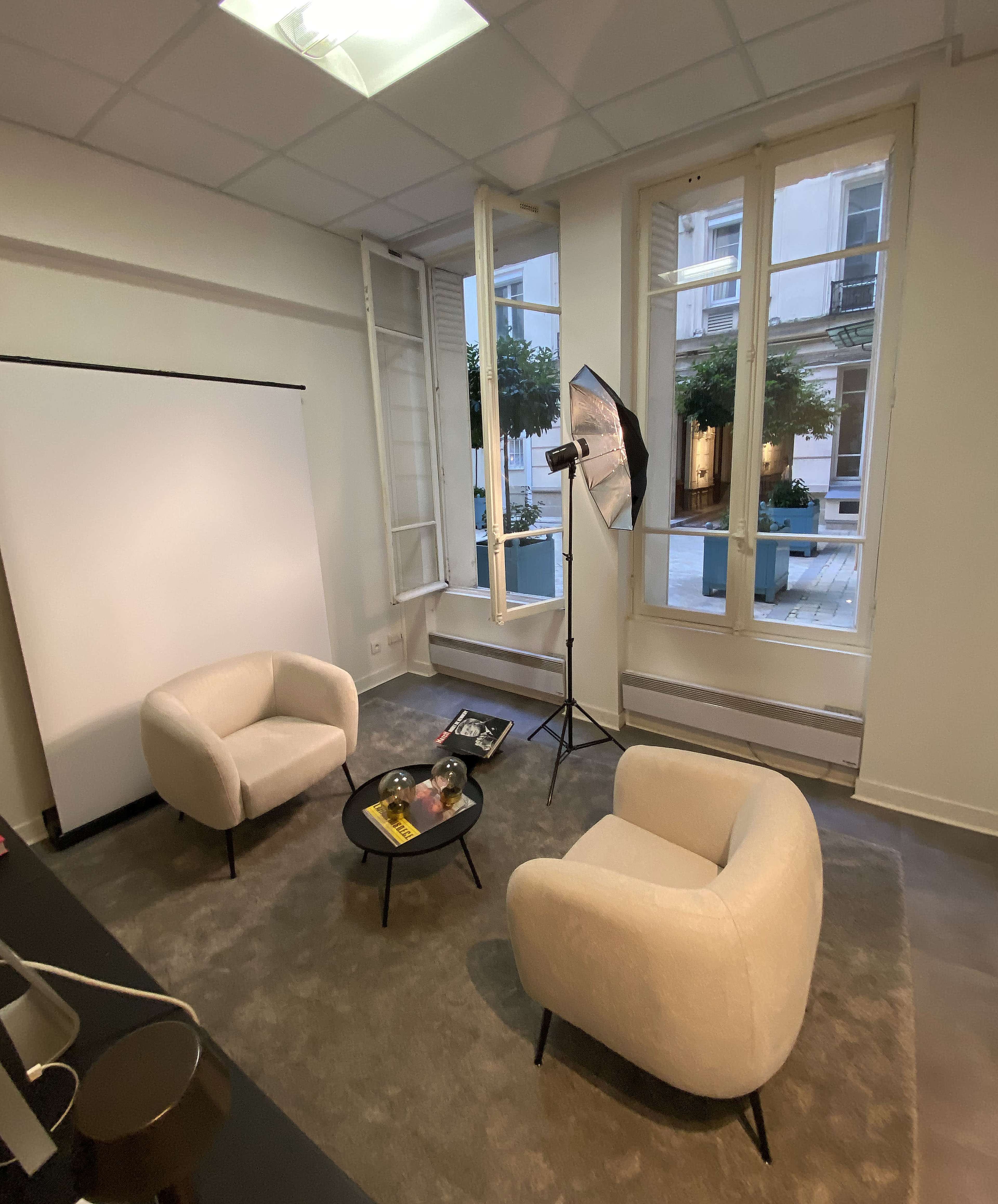 The image shows a minimalist room with two beige armchairs, a small black coffee table, and a backdrop for photo shoots.