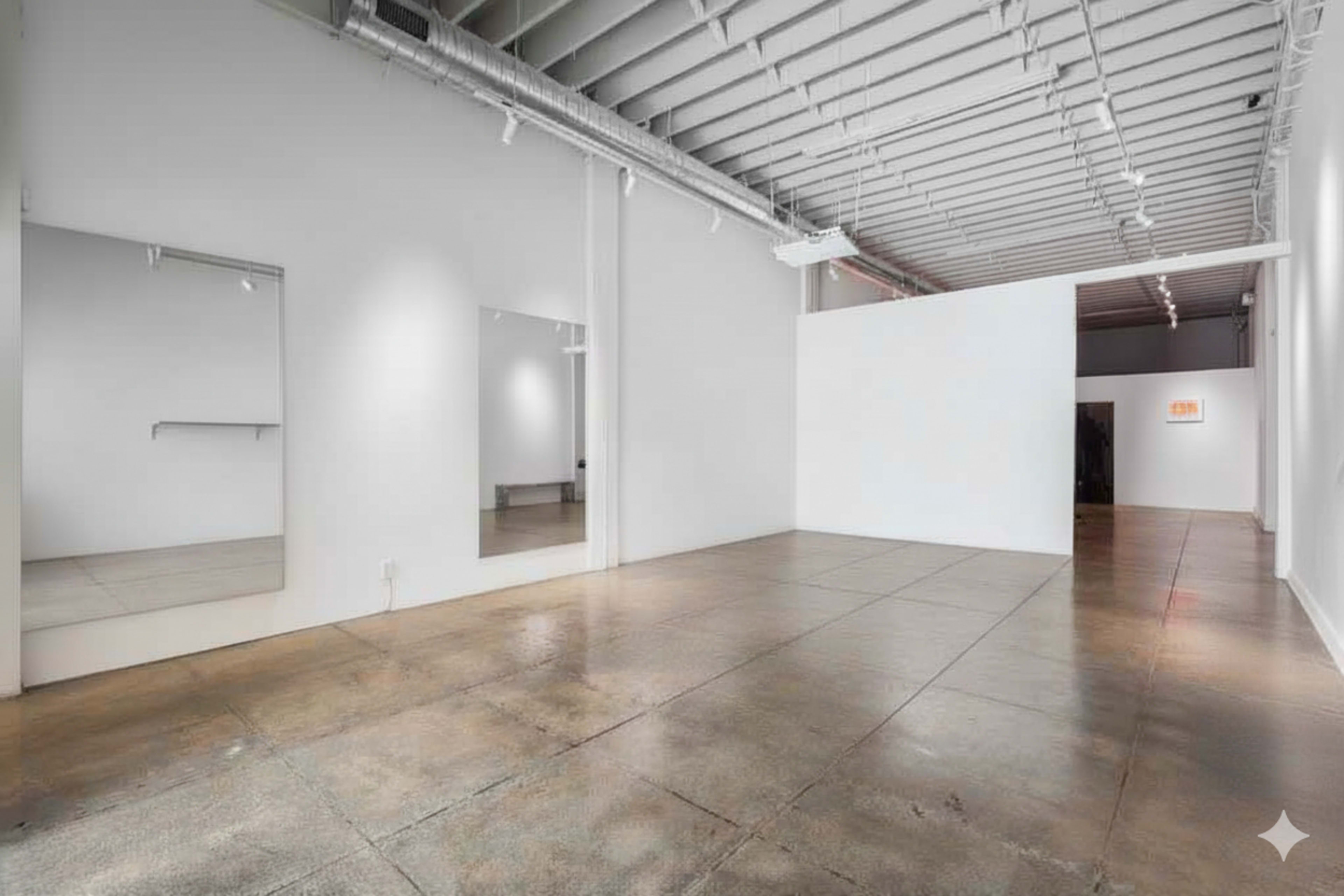 The image shows a spacious, minimalist art gallery with white walls and polished concrete flooring, featuring large mirrors on one wall.