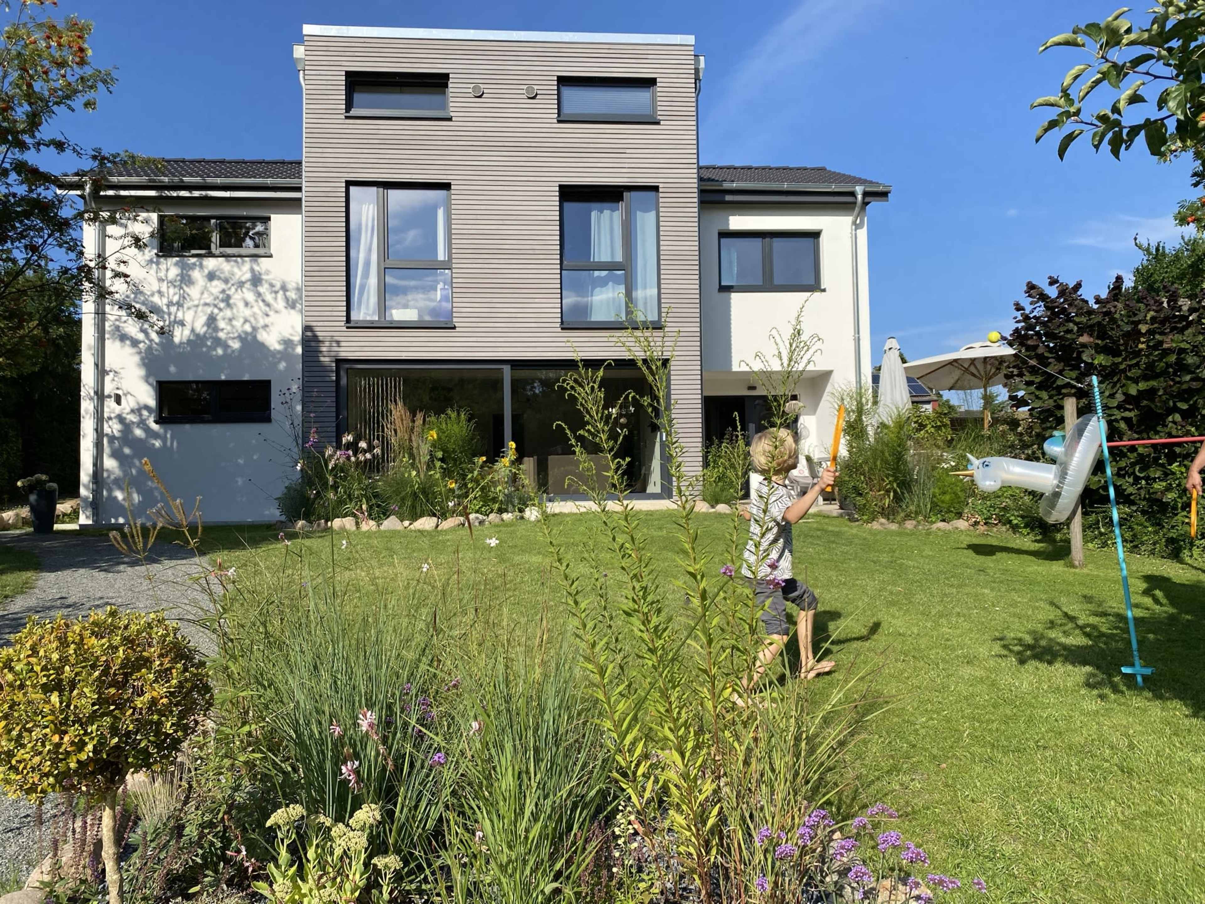 A child plays in the garden of a modern two-story house with large windows and a well-maintained lawn.