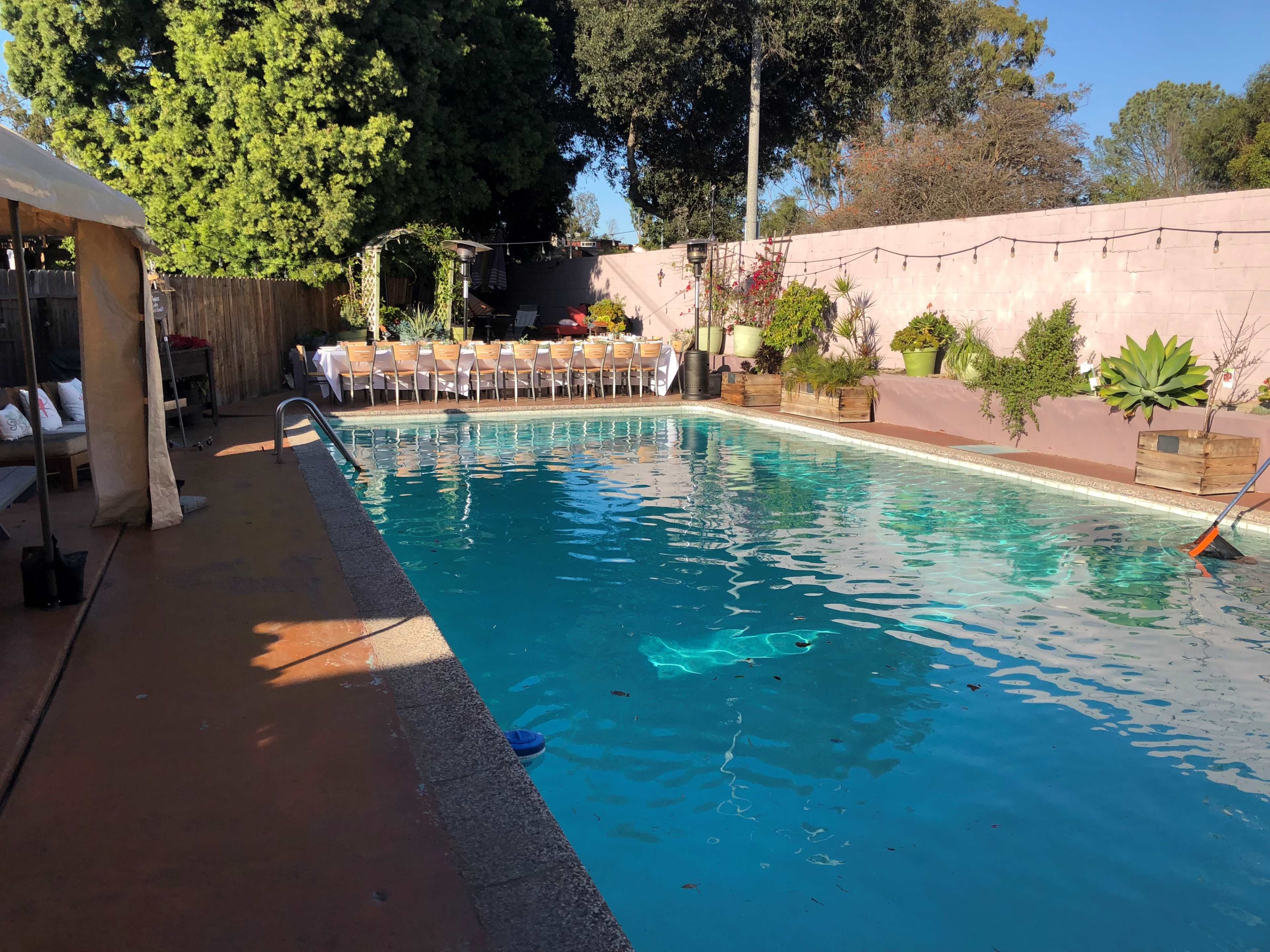 A swimming pool surrounded by a landscaped patio area with lounge chairs and a dining setup under string lights.