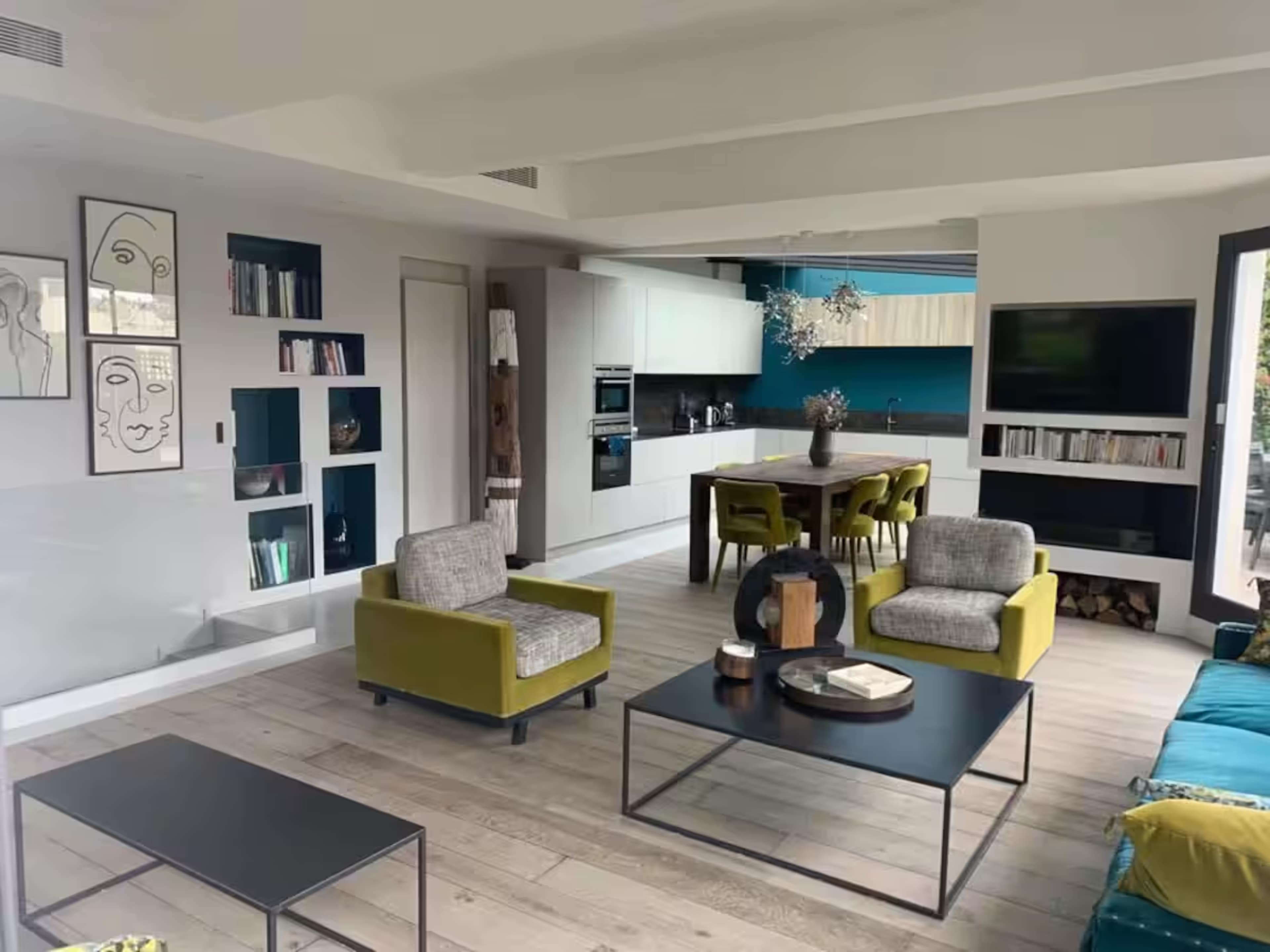 The image shows a modern living space featuring a kitchen area, a dining table, and two armchairs in a light-filled room with wooden flooring and decorative wall art.