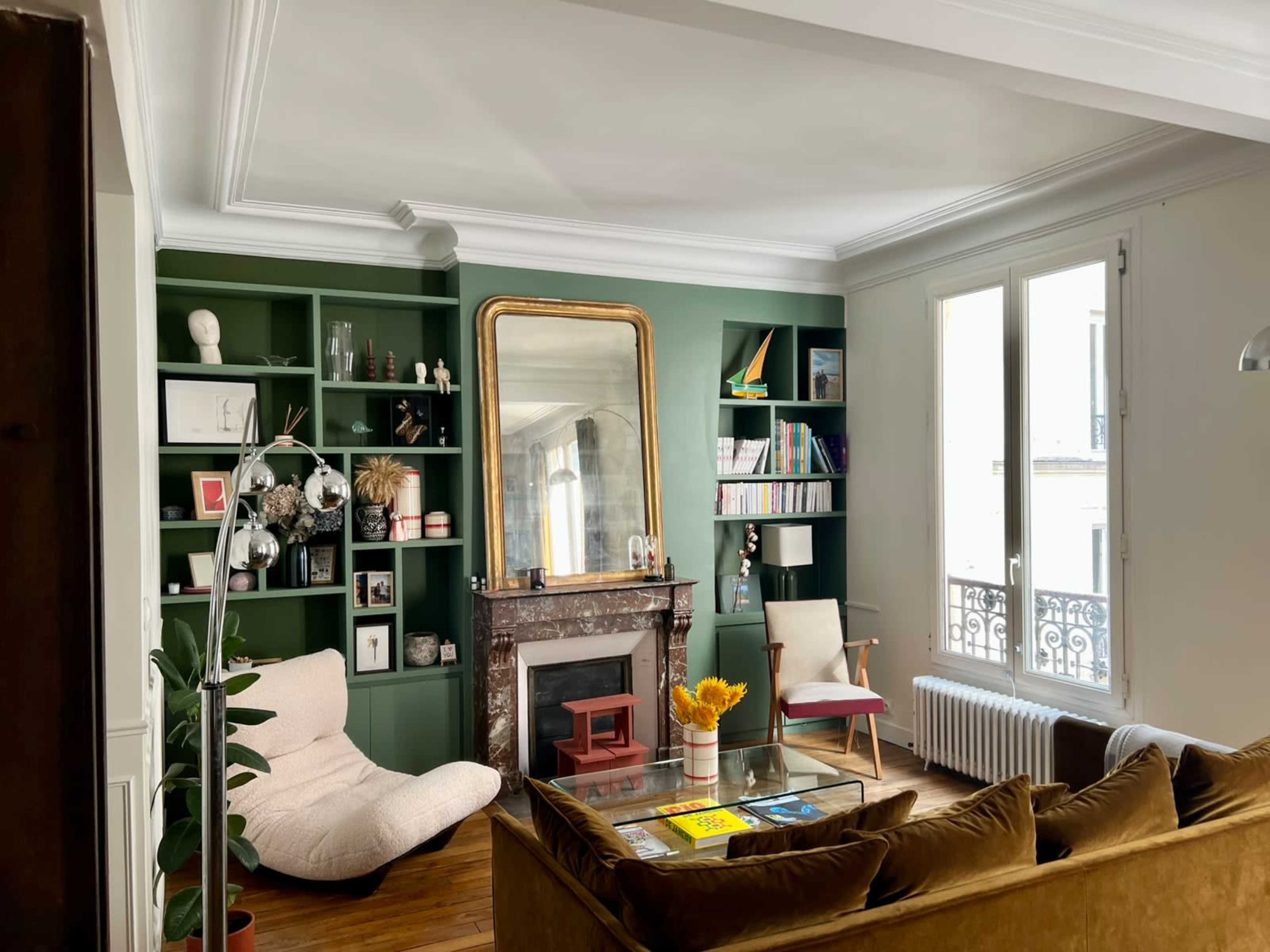 The image depicts a cozy living room featuring a green paneled wall with shelves, a large mirror, and a mix of modern and vintage furnishings, including a light-colored armchair and a brown sofa.